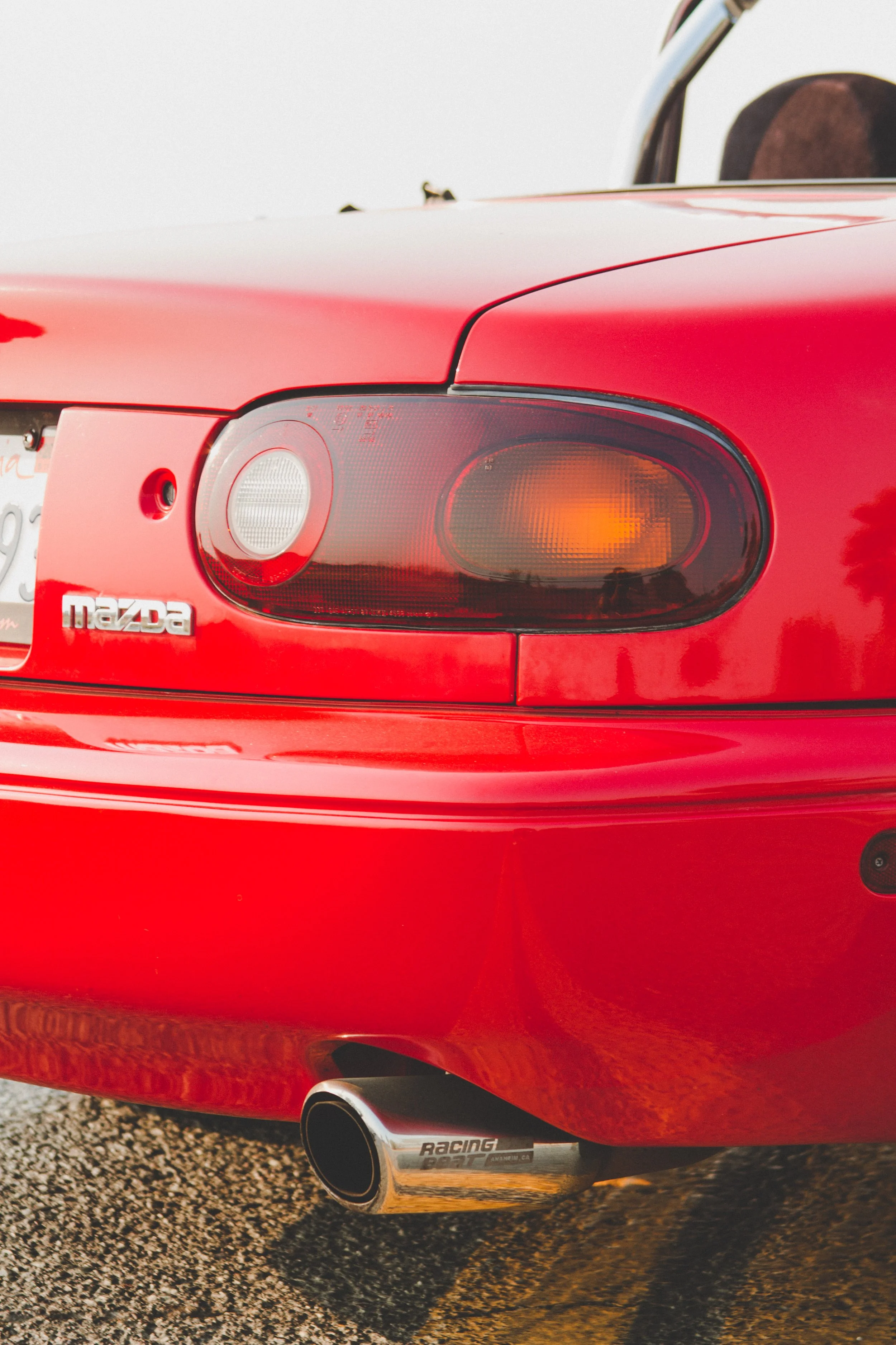 Close-up of the rear of a red Mazda sports car, showing tail lights, a Mazda badge, and an aftermarket racing exhaust pipe.
