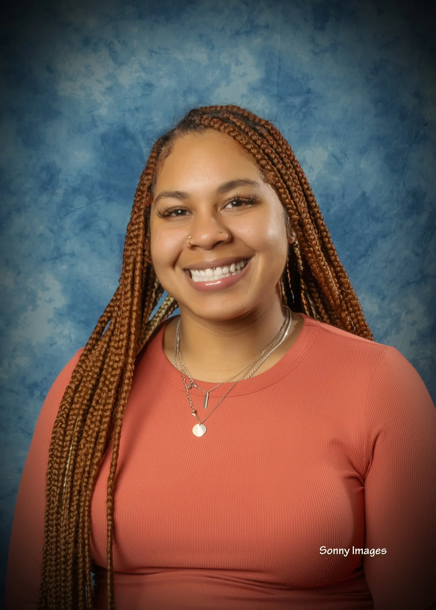 Young woman with long, brown braided hair smiling against a blue textured background, wearing a coral long-sleeve top and layered necklaces.