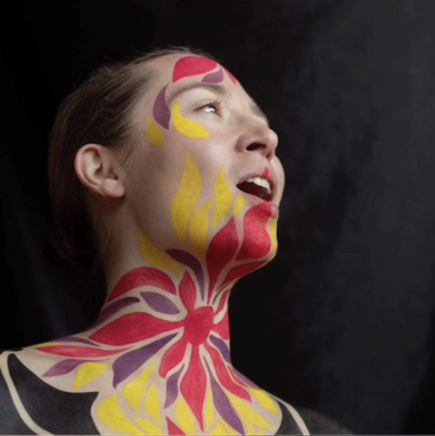 A woman with face and neck painted in a floral pattern using red, yellow, and purple colors, looking up and to the side against a dark background.