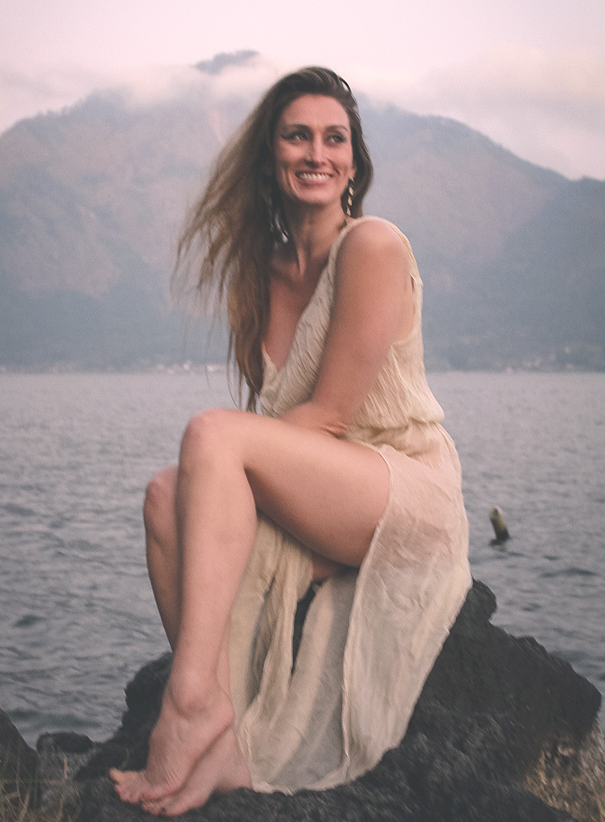 A woman with long hair wearing a white tunic sitting on a large rock by the water, smiling with mountains in the background during sunset.