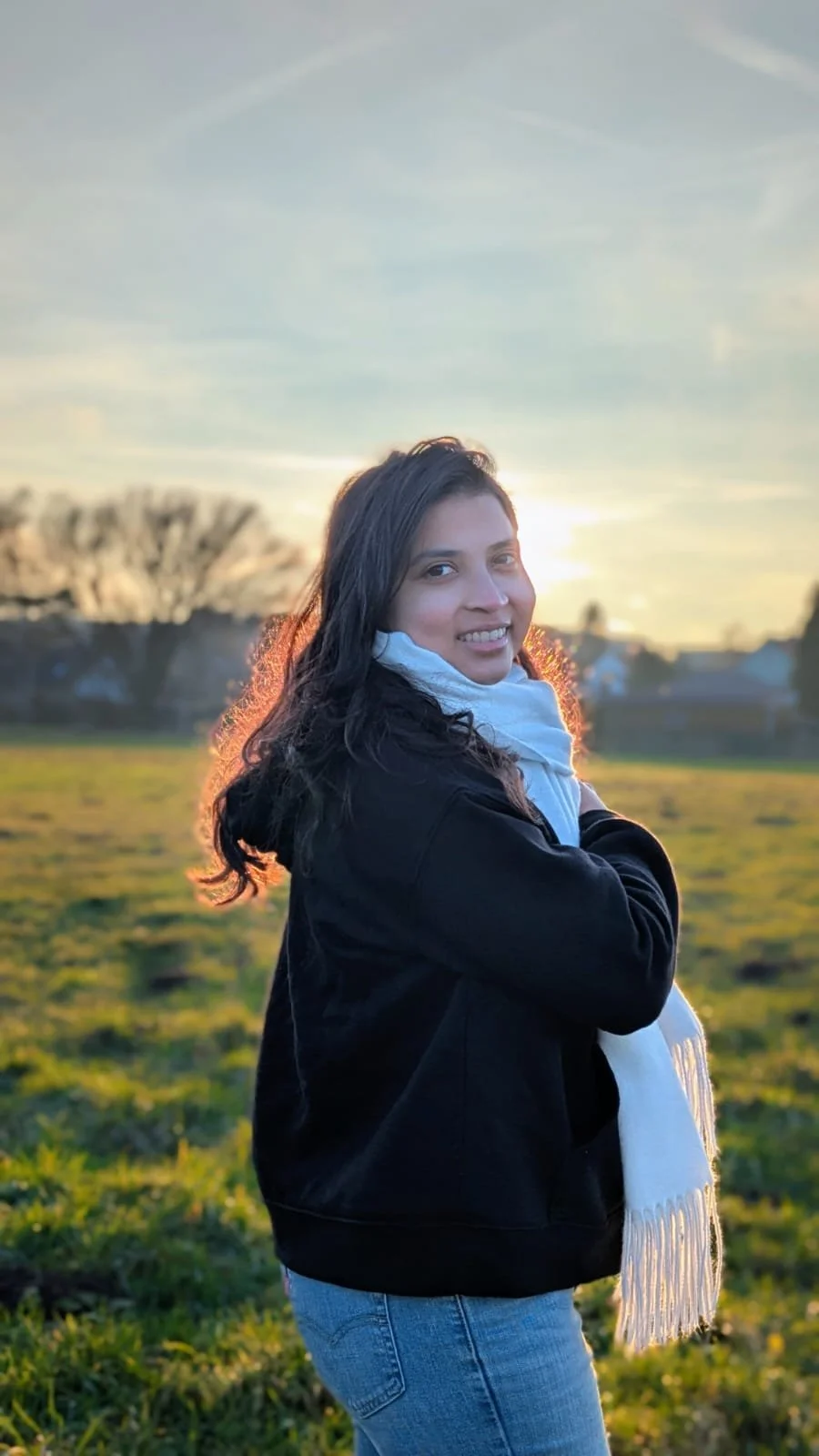 A woman with long dark hair wearing a black jacket and white scarf stands outdoors during sunset, smiling at the camera with a grassy field and trees in the background.