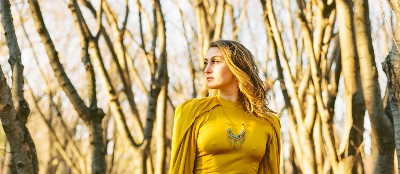 A woman with wavy hair wearing a yellow dress and necklace, standing among trees with sunlight filtering through during autumn.