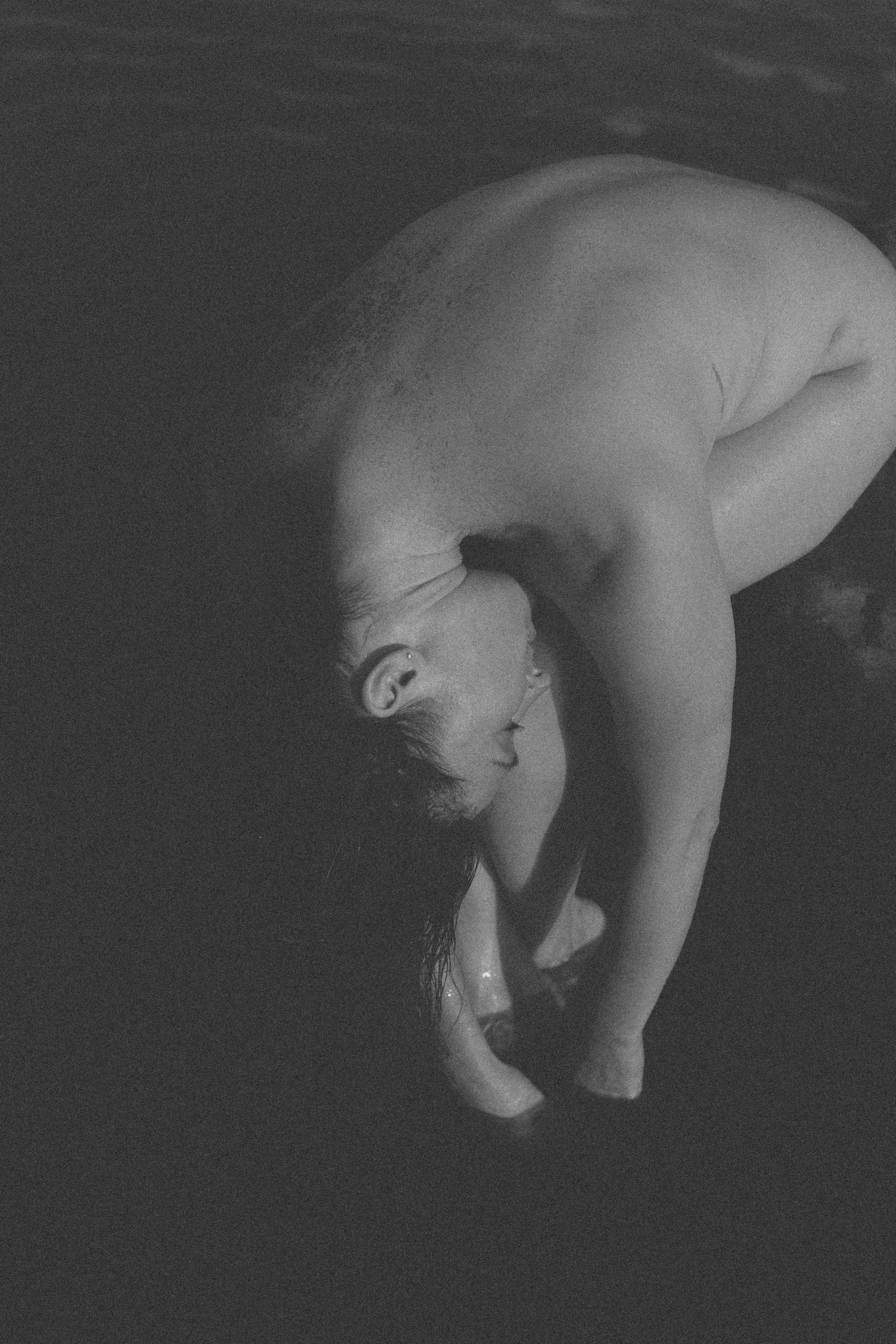 Artful nude image of a woman with cascading hair reaching downwards toward water her feet and hands melt into,. She is curled up over herself as she sits on a rock.