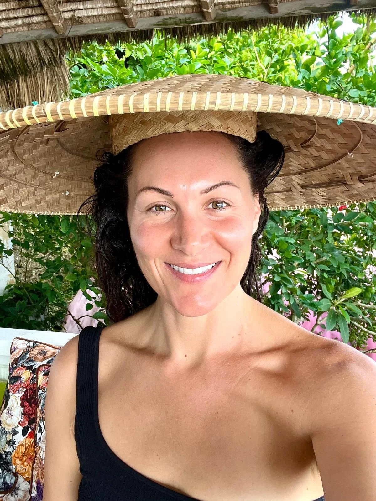 Woman with dark curly hair and light skin, smiling, wearing a black top and a large straw sun hat, in a lush green outdoor setting.