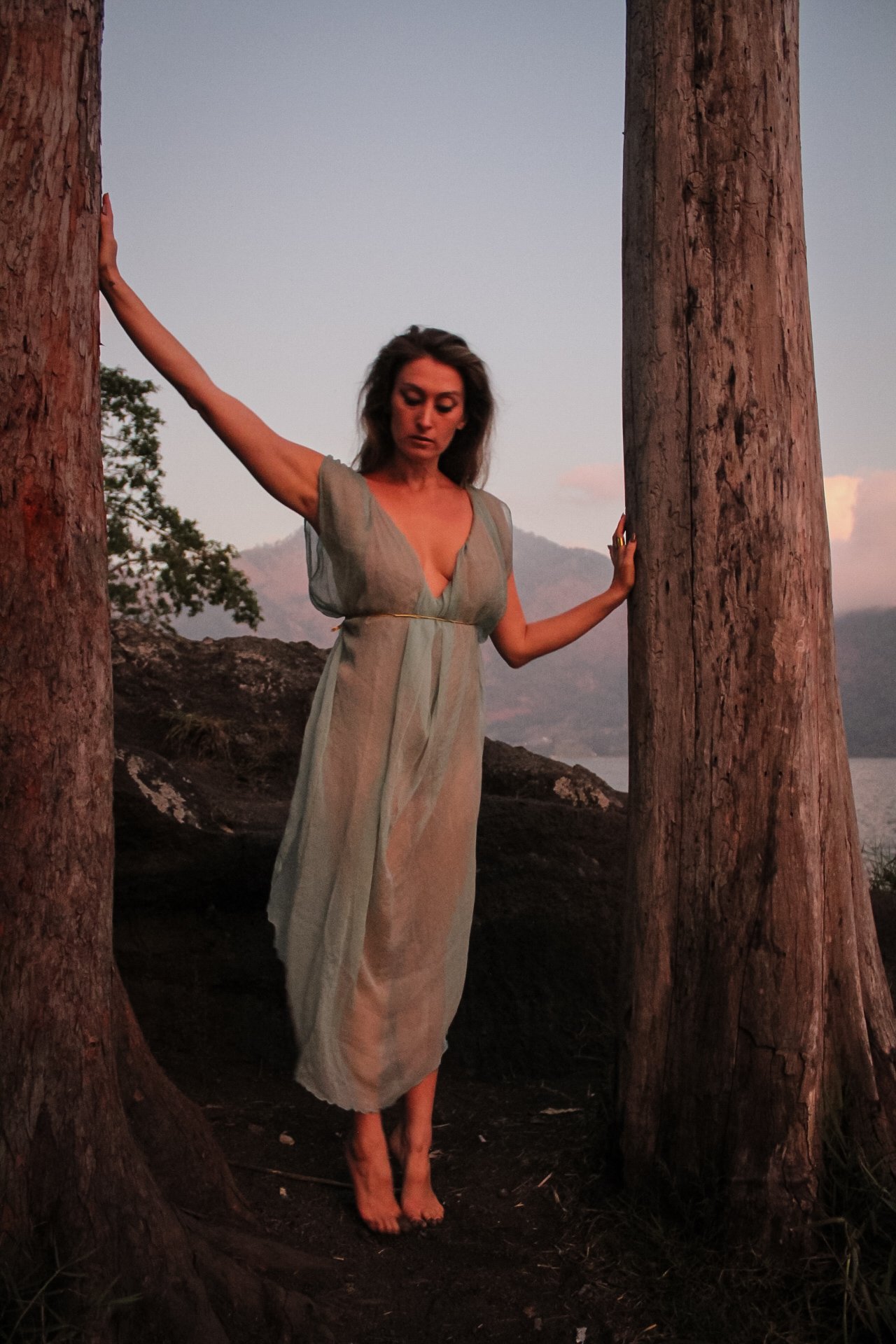 A woman in a sheer, blue tunic standing barefoot between two large trees outdoors during sunset, with distant mountains and a body of water in the background.