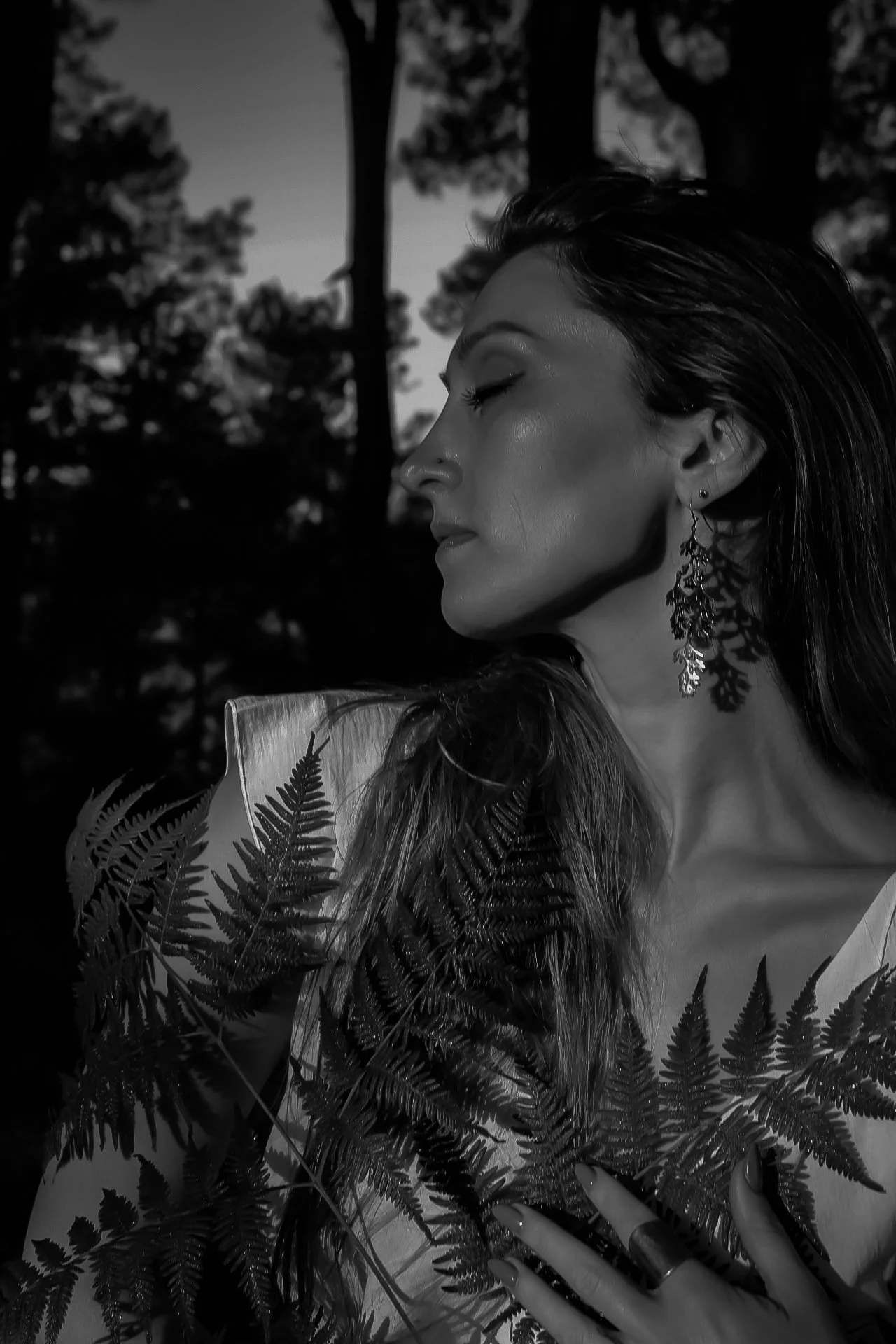 A woman with long hair, earrings, and makeup, appears to be enjoying nature with ferns on her shoulder and chest, set against a backdrop of trees and sky in a black-and-white photograph.