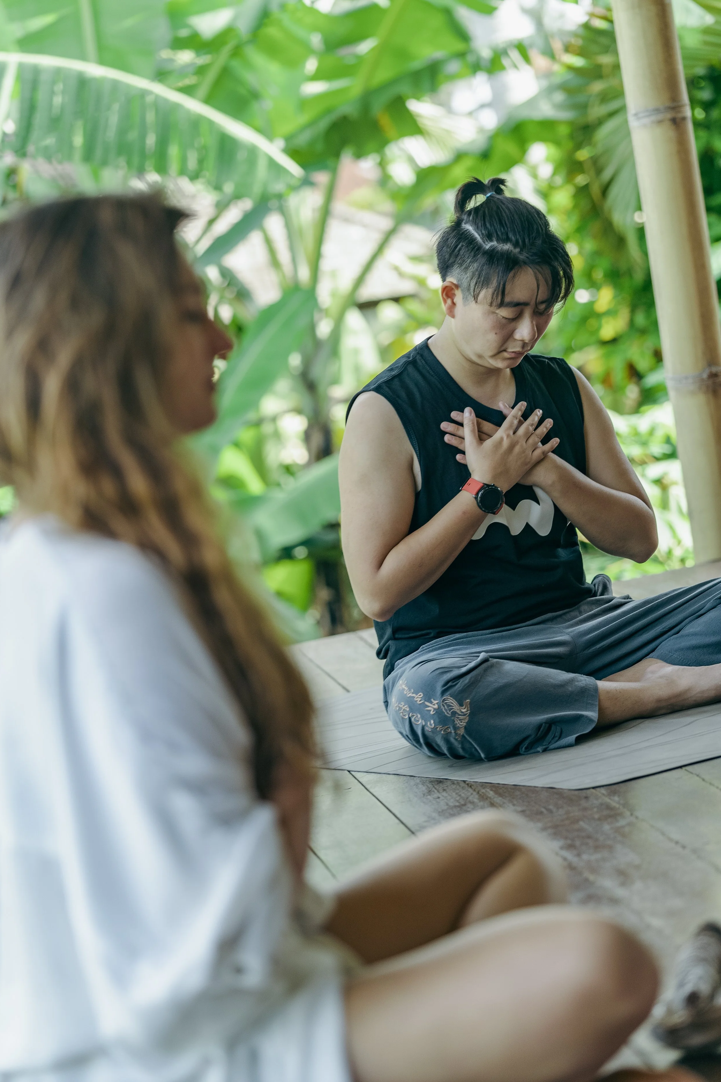 Two people sitting cross-legged on a mat outdoors, with one person meditating with hands on chest and eyes closed, in a lush green environment.