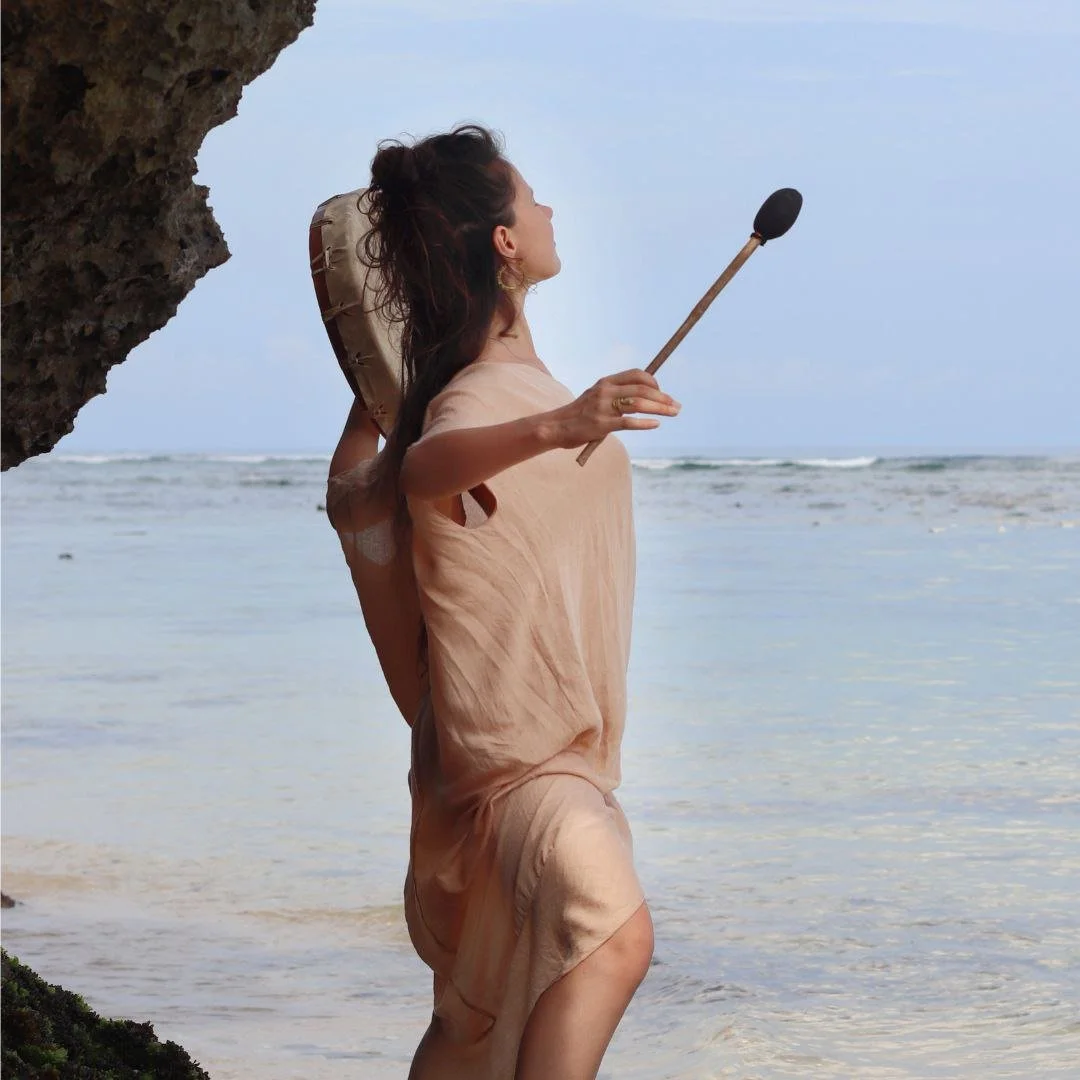 A woman on the beach holding a mallet, facing towards the ocean, with a large rock over her shoulder.