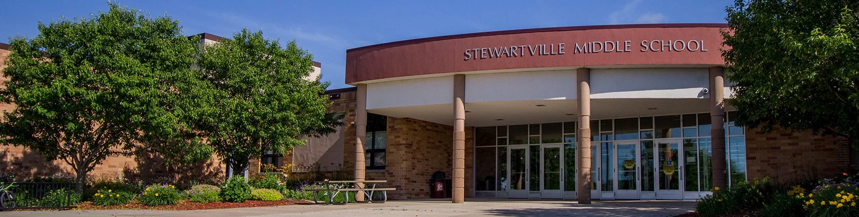 Front view of Stewartville Middle School with trees, flower beds, and a clear blue sky.