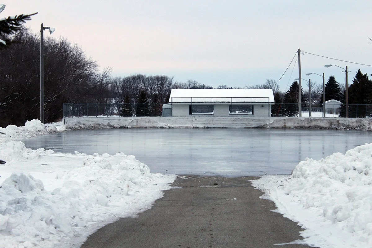 An outdoor ice skating rink surrounded by snow with a building in the background and trees on either side.