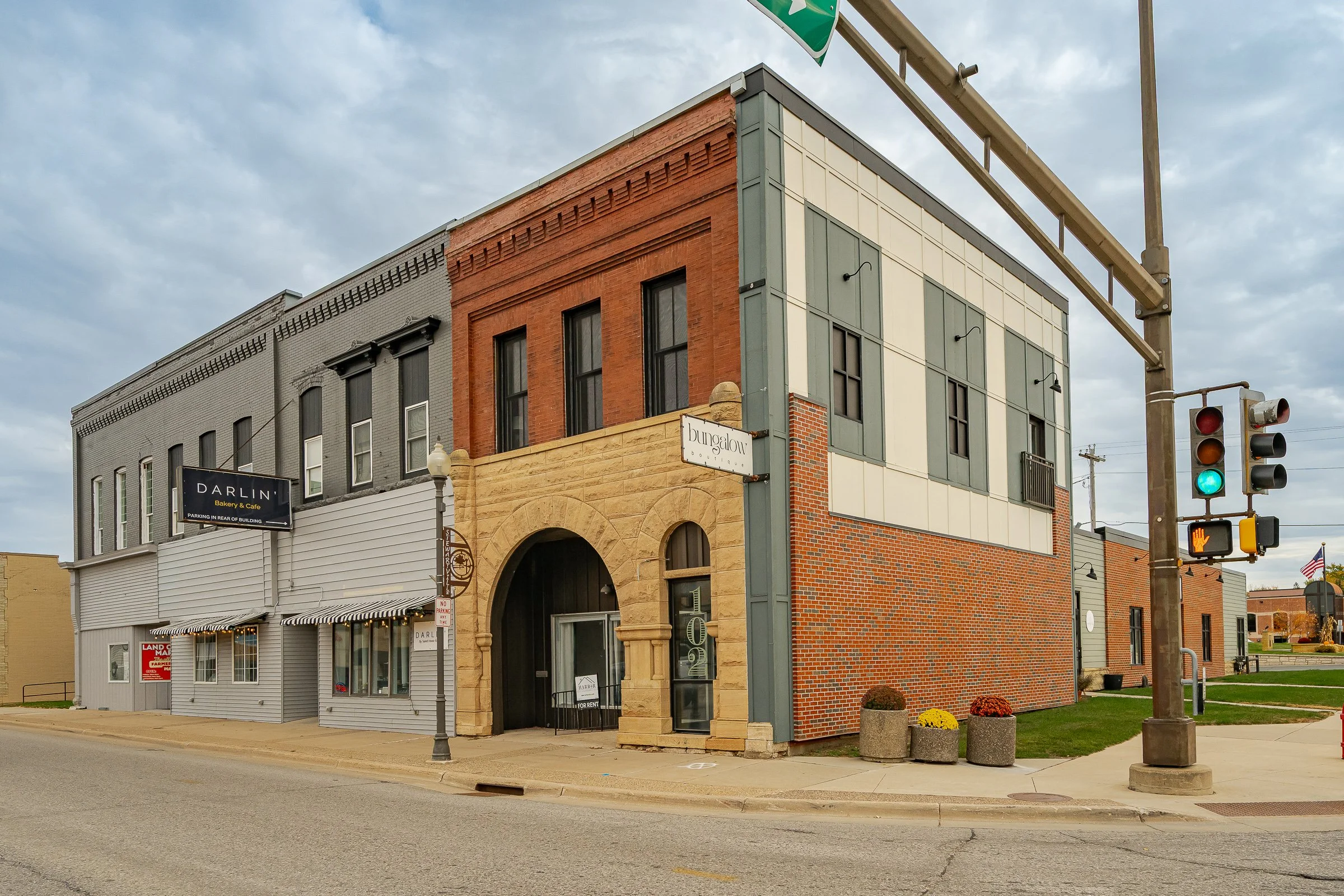 A street corner with a colorful, multi-story building featuring a mix of brick and painted facades, a black and white sign for Darlin' Bakery & Cafe, a 'For Rent' sign in a window, street lamps, concrete planters, and a traffic light showing green, under a partly cloudy sky.