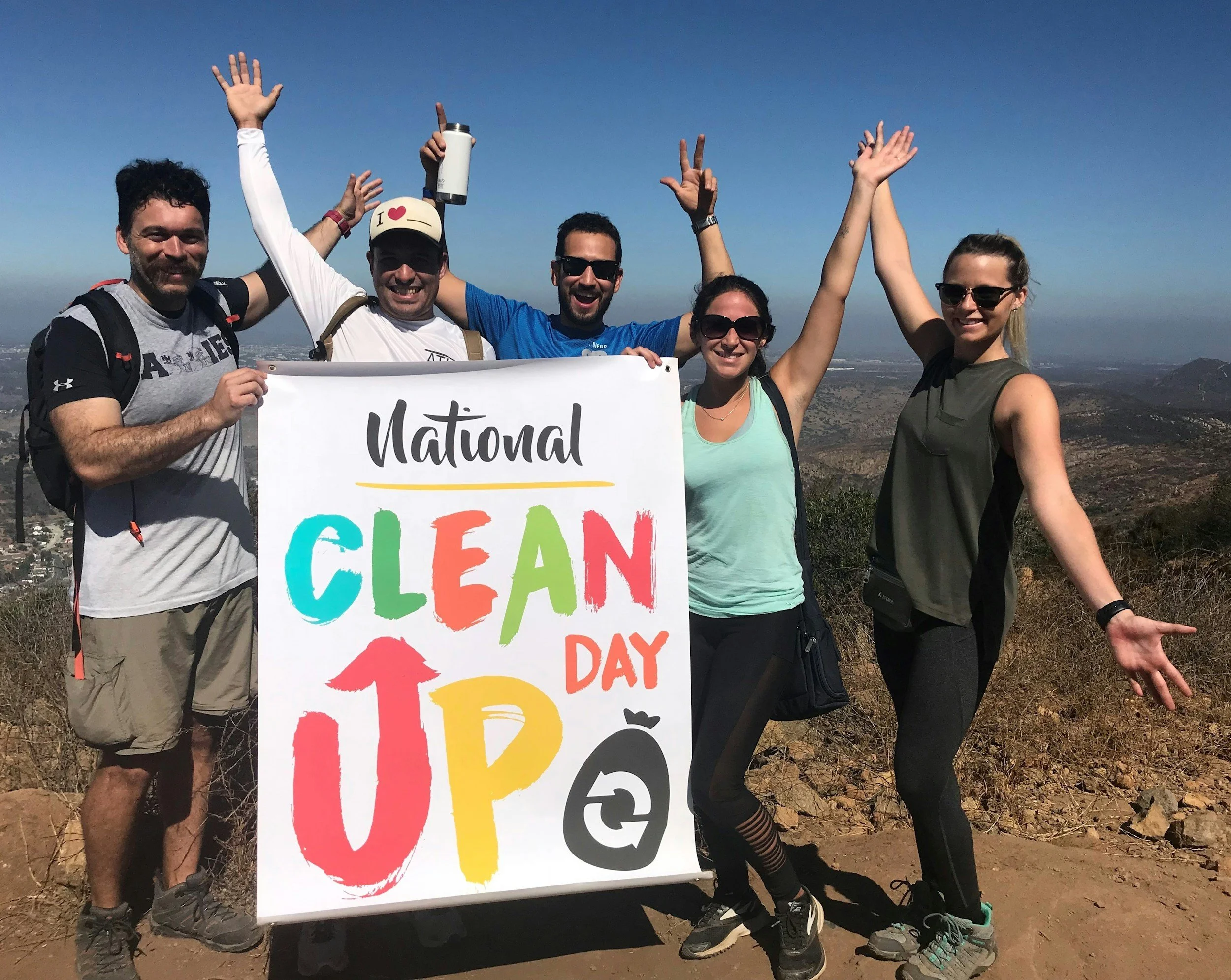 A group of five hikers holding a colorful sign that reads 'National Clean Up Day' on a mountain trail with scenic landscape and blue sky in the background, celebrating environmental effort.