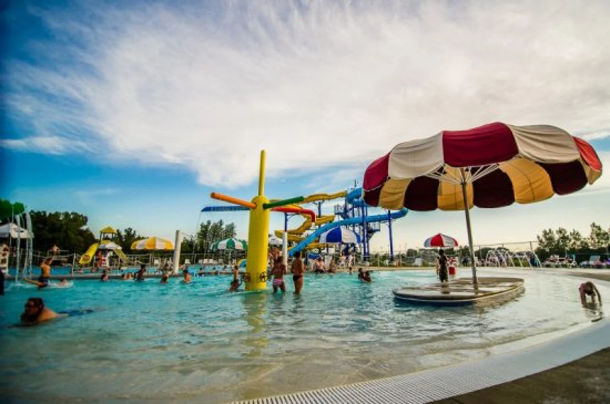 A lively outdoor water park with multiple water slides, a large striped umbrella over a shallow pool, and many people enjoying the water on a bright day with a partly cloudy sky.