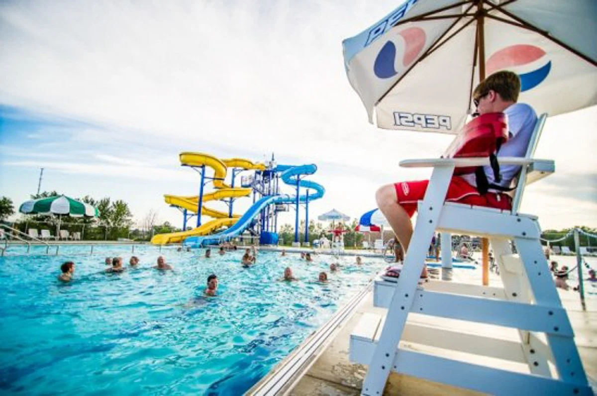 A man relaxing on a lifeguard chair under a Pepsi umbrella beside a swimming pool with people swimming and water slides in the background on a sunny day.
