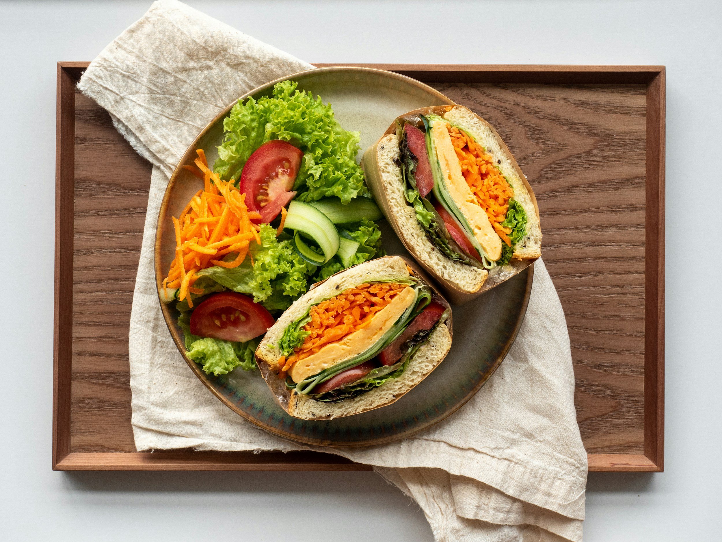 A plate with a cut sandwich showing layers of lettuce, tomato, shredded carrots, cucumber, and egg salad, served alongside a side salad of lettuce, tomato, shredded carrots, and cucumber slices on a wooden tray.