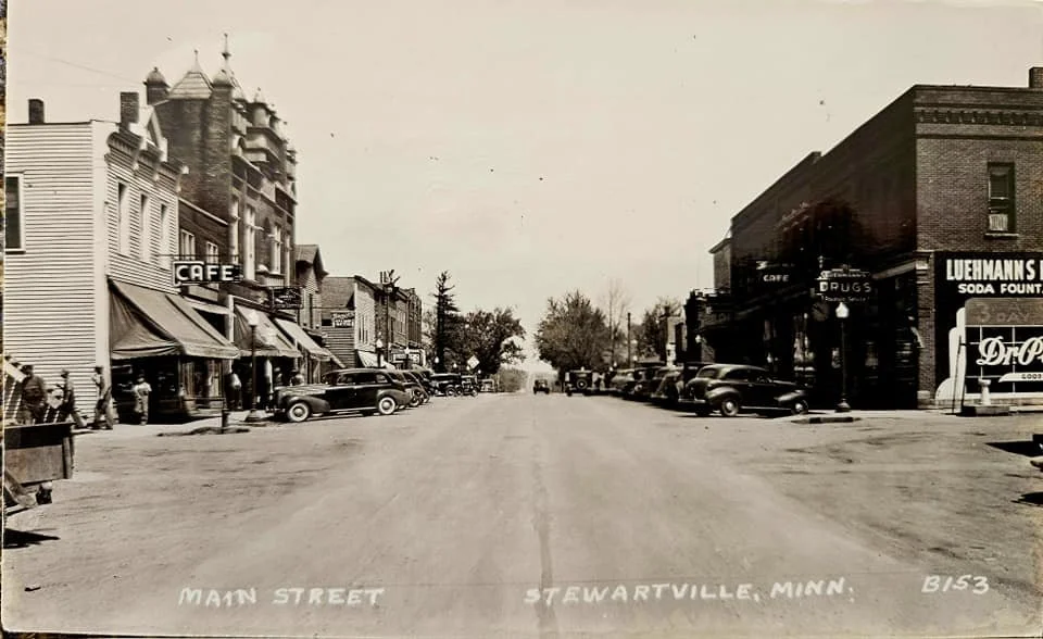 Black and white photo of Main Street in Stewartville, Minnesota, showing historic buildings, parked cars, storefronts including a cafe and drug store, and a soda fountain sign.