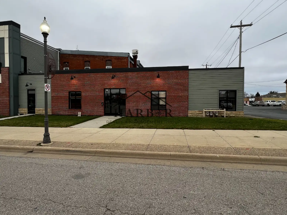 A modern commercial building with a brick facade and gray siding, situated on a sidewalk in a small town or suburban area, under an overcast sky.