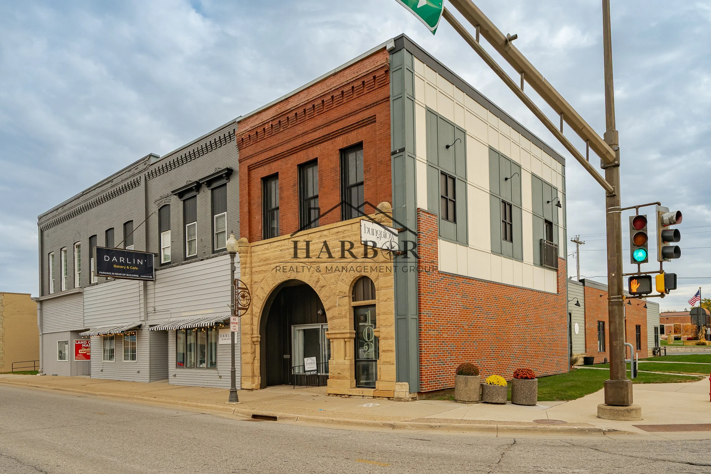 A street corner with a colorful, multi-story building featuring a mix of brick and painted facades, a black and white sign for Darlin' Bakery & Cafe, a 'For Rent' sign in a window, street lamps, concrete planters, and a traffic light showing green, under a partly cloudy sky.