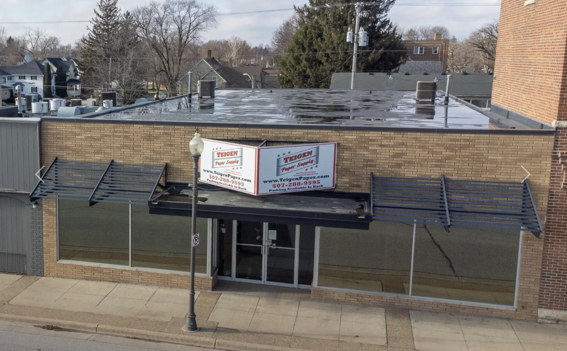 Empty commercial storefront with large glass windows, a brick facade, and a black metal awning. A sign on the building advertises Teigen Paper Supply. A street lamp is in front of the store, and there is a wet, reflective roof on top of the building.