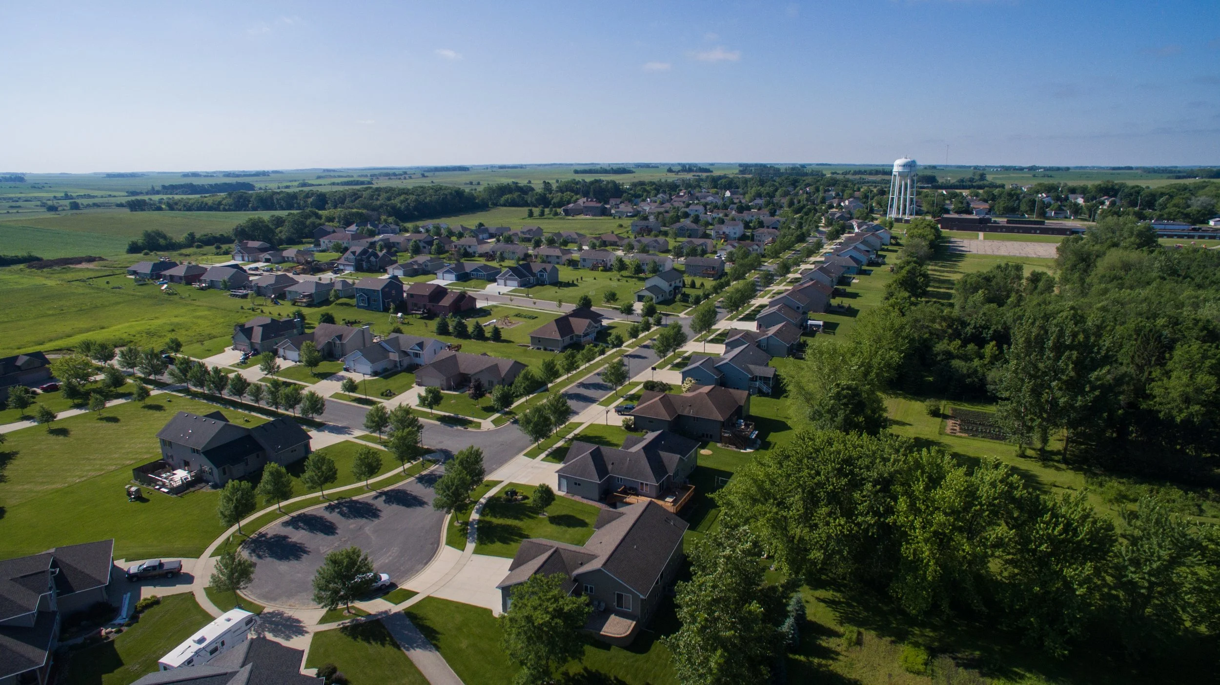 Aerial view of a suburban neighborhood with houses, trees, roads, and a water tower in the distance under a blue sky.