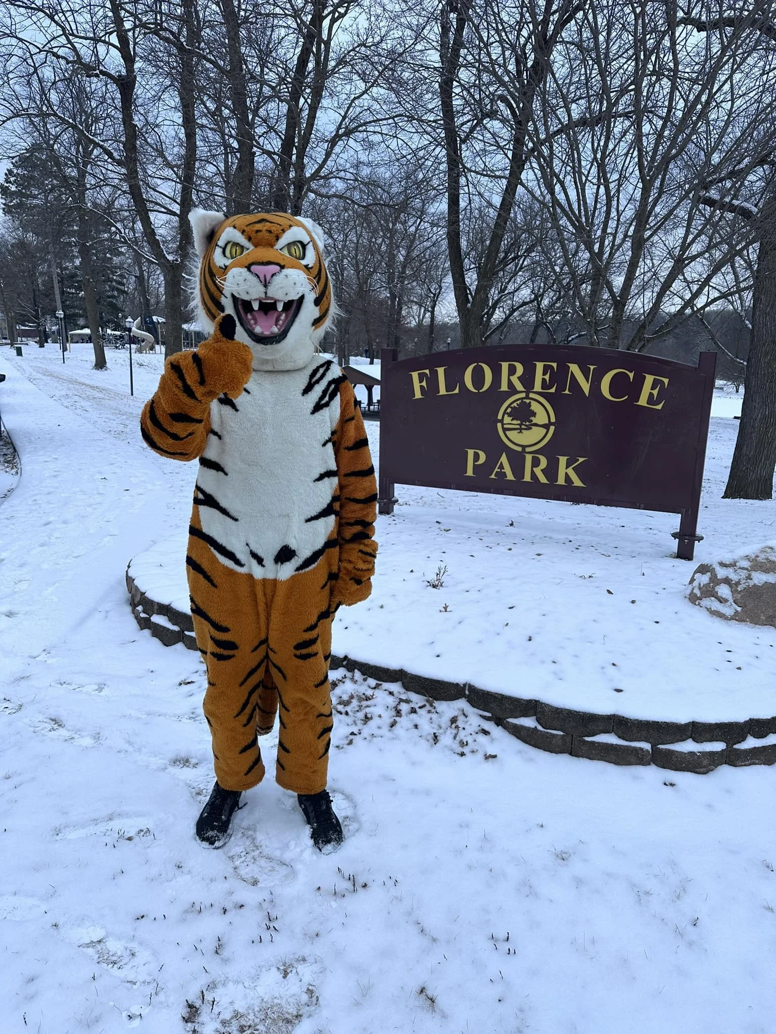 Person in a tiger mascot costume standing on snow at Florence Park, pointing upward, with trees and park sign in the background.