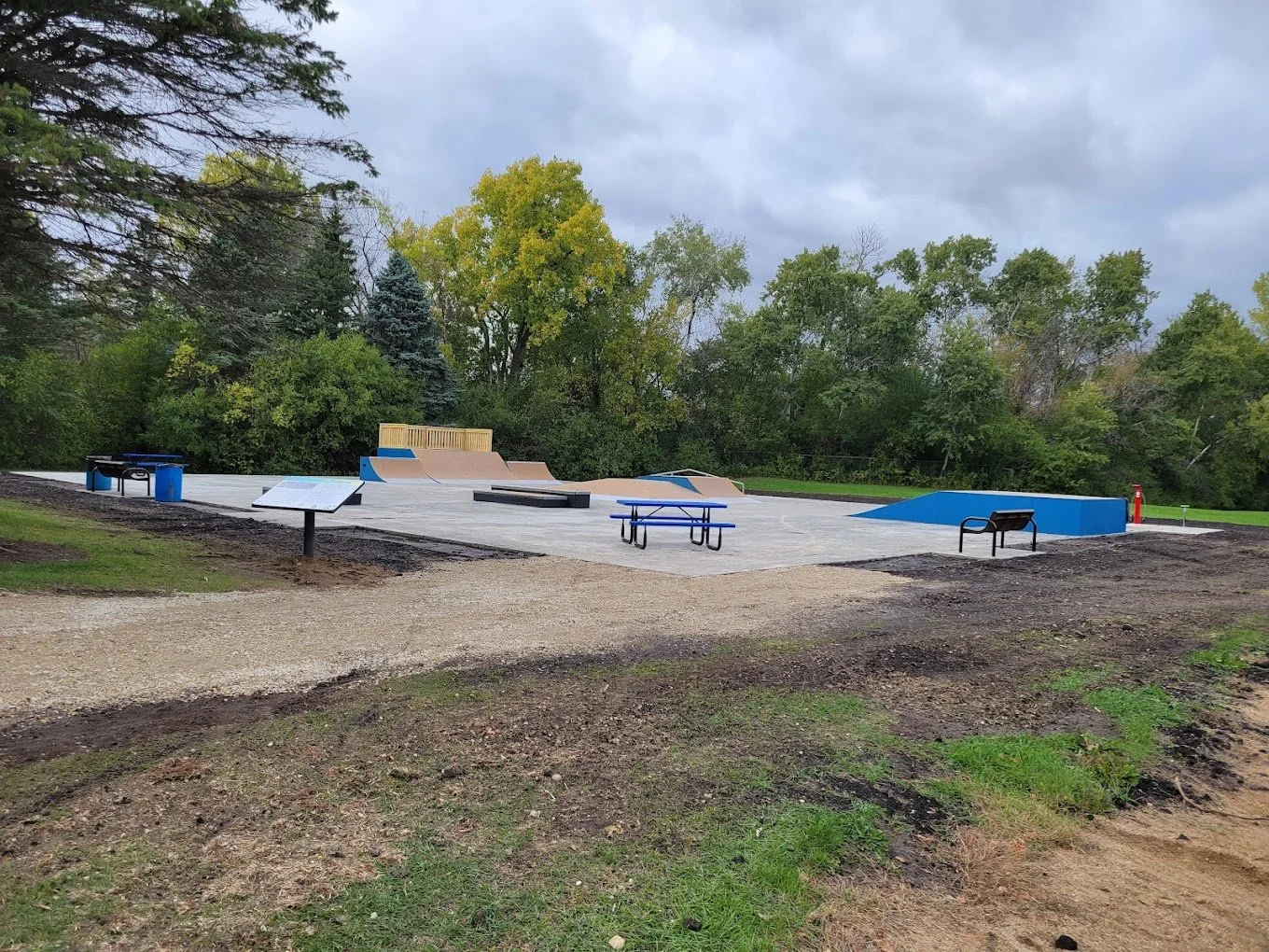 A skatepark with multiple ramps and rails, surrounded by trees and cloudy sky, with some dirt and grass in the foreground.
