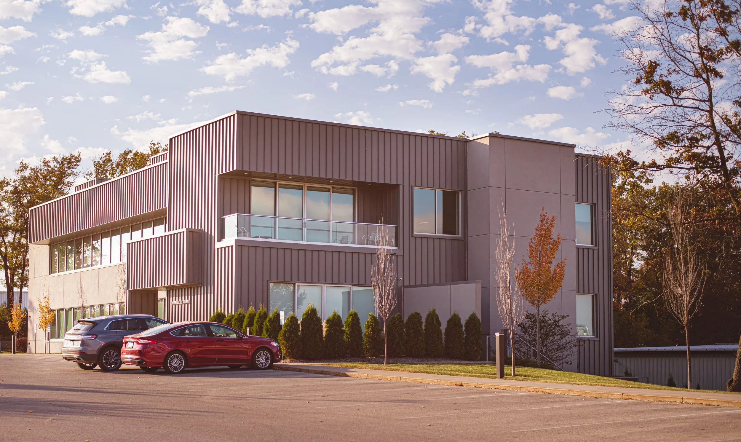 Modern office building with large windows, a balcony, landscaped area with trees and shrubs, and parked cars in the foreground under a partly cloudy sky.