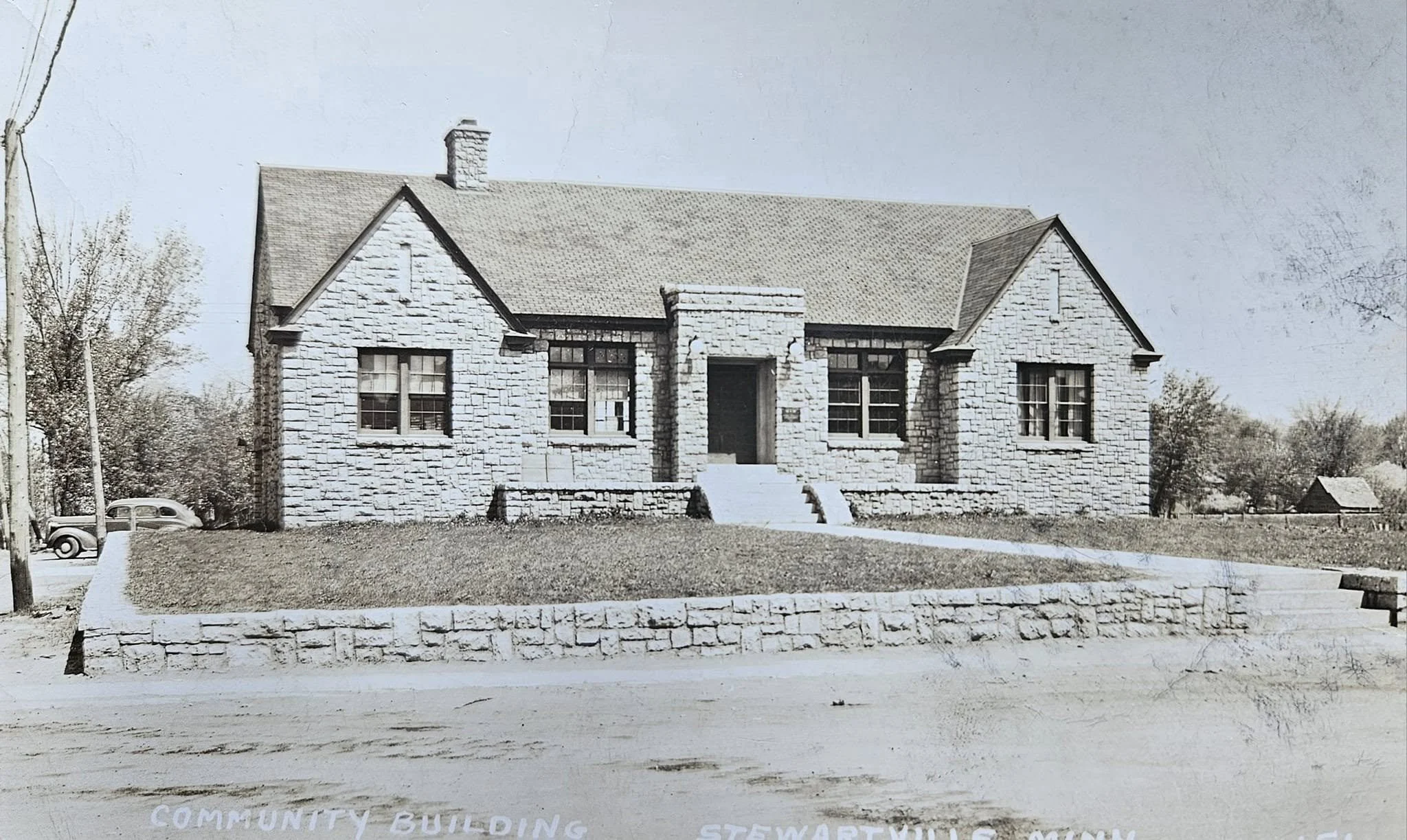 Black and white photo of a stone community building with steps leading to the front door, surrounded by trees and a small yard.
