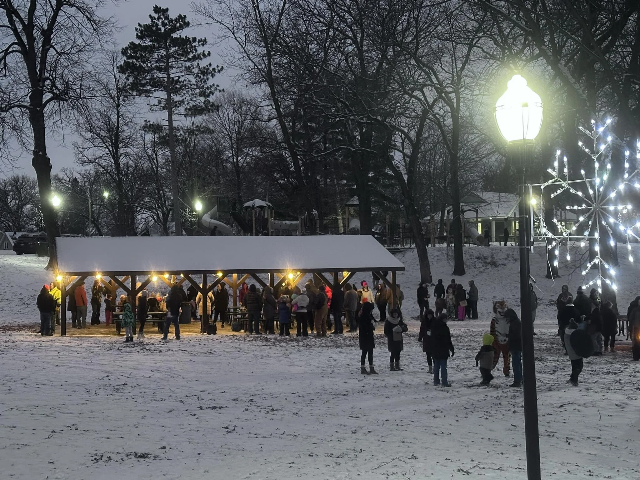 People gathering at a park pavilion decorated with lights during winter, with snow on the ground and trees, and a playground in the background.