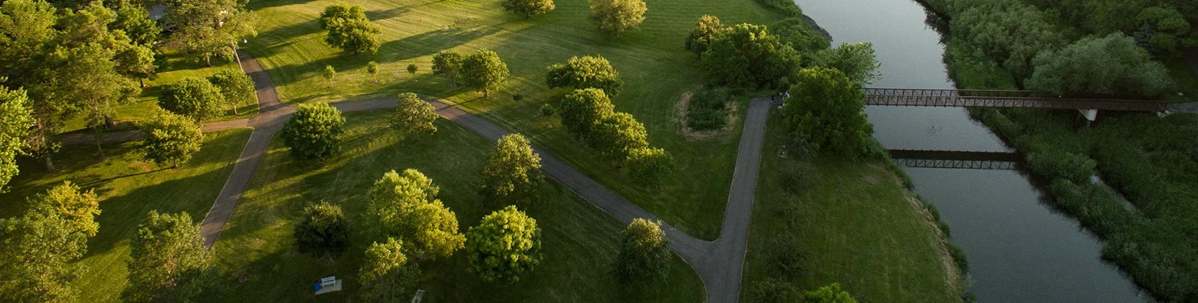 Aerial view of a park with winding paths, green trees, and a waterbody with a bridge