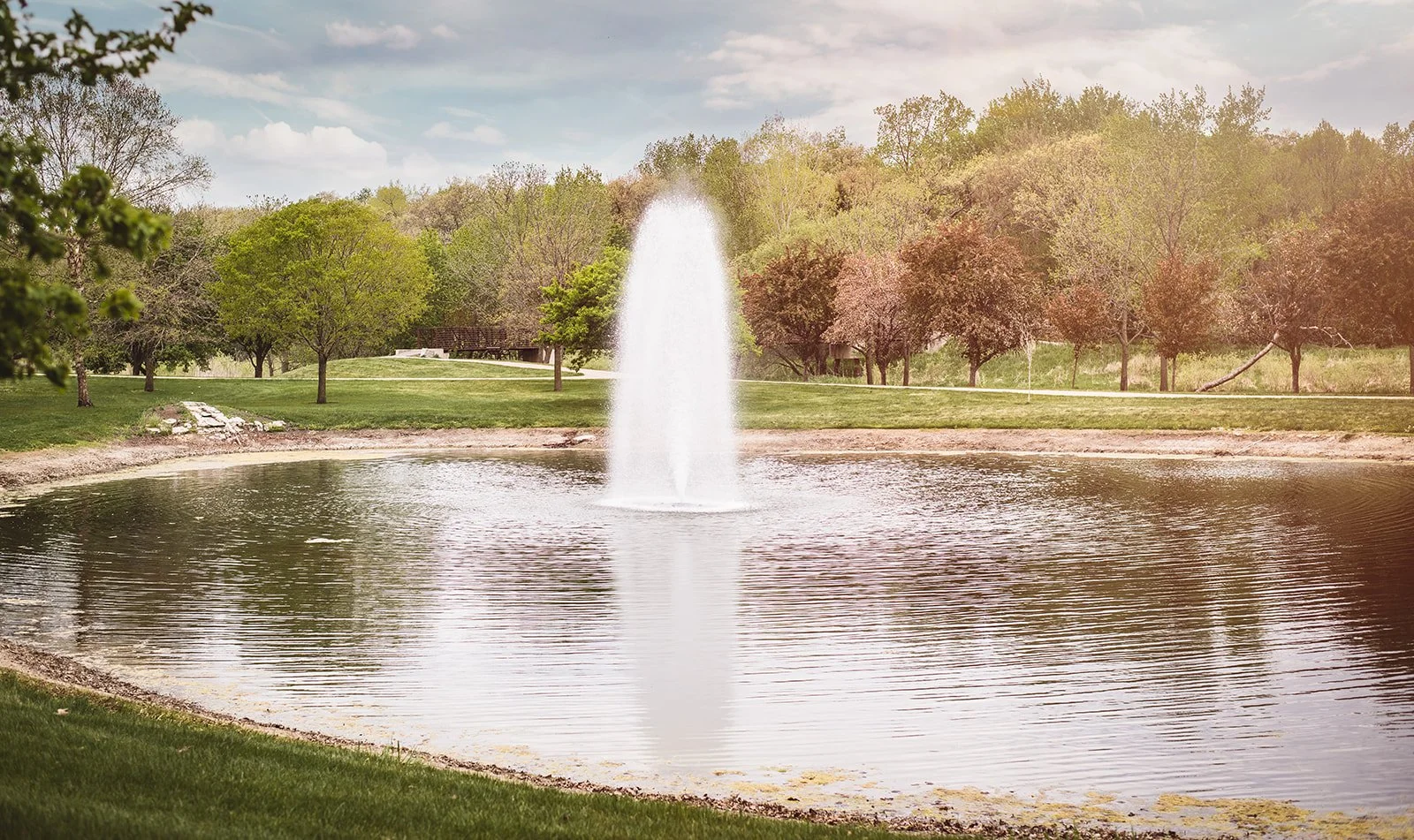 A peaceful park scene with a central water fountain, surrounded by green grass, trees, and a cloudy sky.