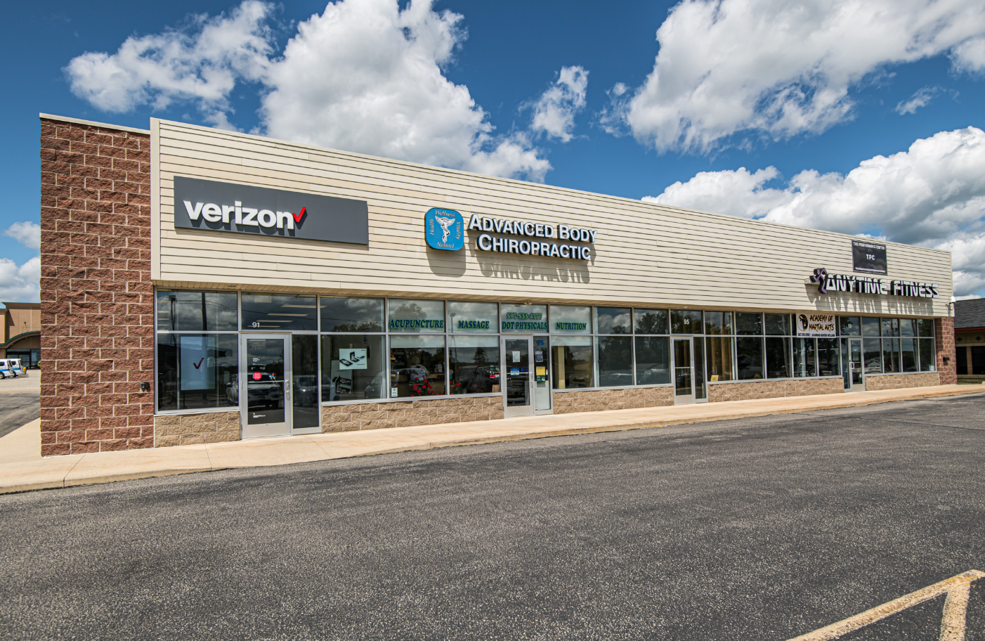 A strip mall storefront featuring businesses such as Verizon, Advanced Body Chiropractic, City Time Fitness, and an acupuncture, massage, and nutrition clinic, with a parking lot in front and a partly cloudy sky above.