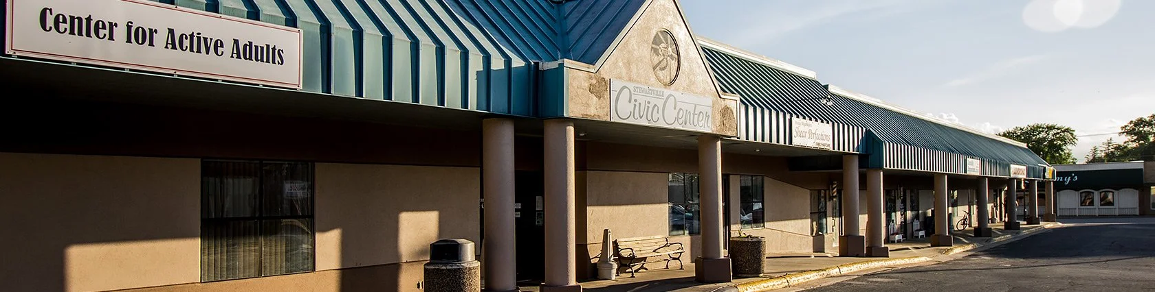 A strip mall with storefronts under a blue metal awning, including a sign for 'Center for Active Adults' and another for 'Civic Center', with a parking lot in front and a bicycle visible at one shop.