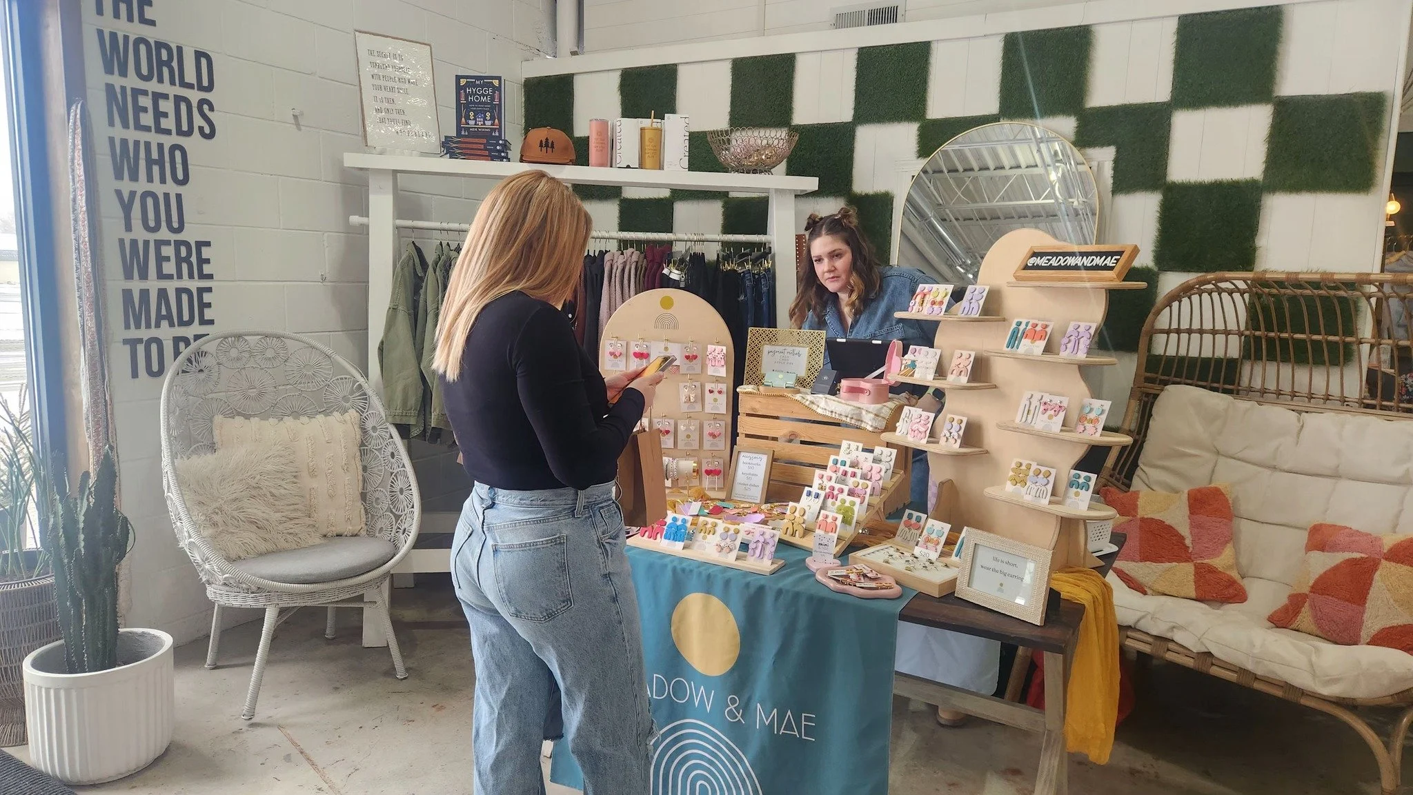 A woman browsing handmade jewelry at a small boutique stand, while a store clerk stands behind the display. The boutique has a display with jewelry pieces, a cozy seating area with a rattan sofa and cushions, and decorative wall elements including a 