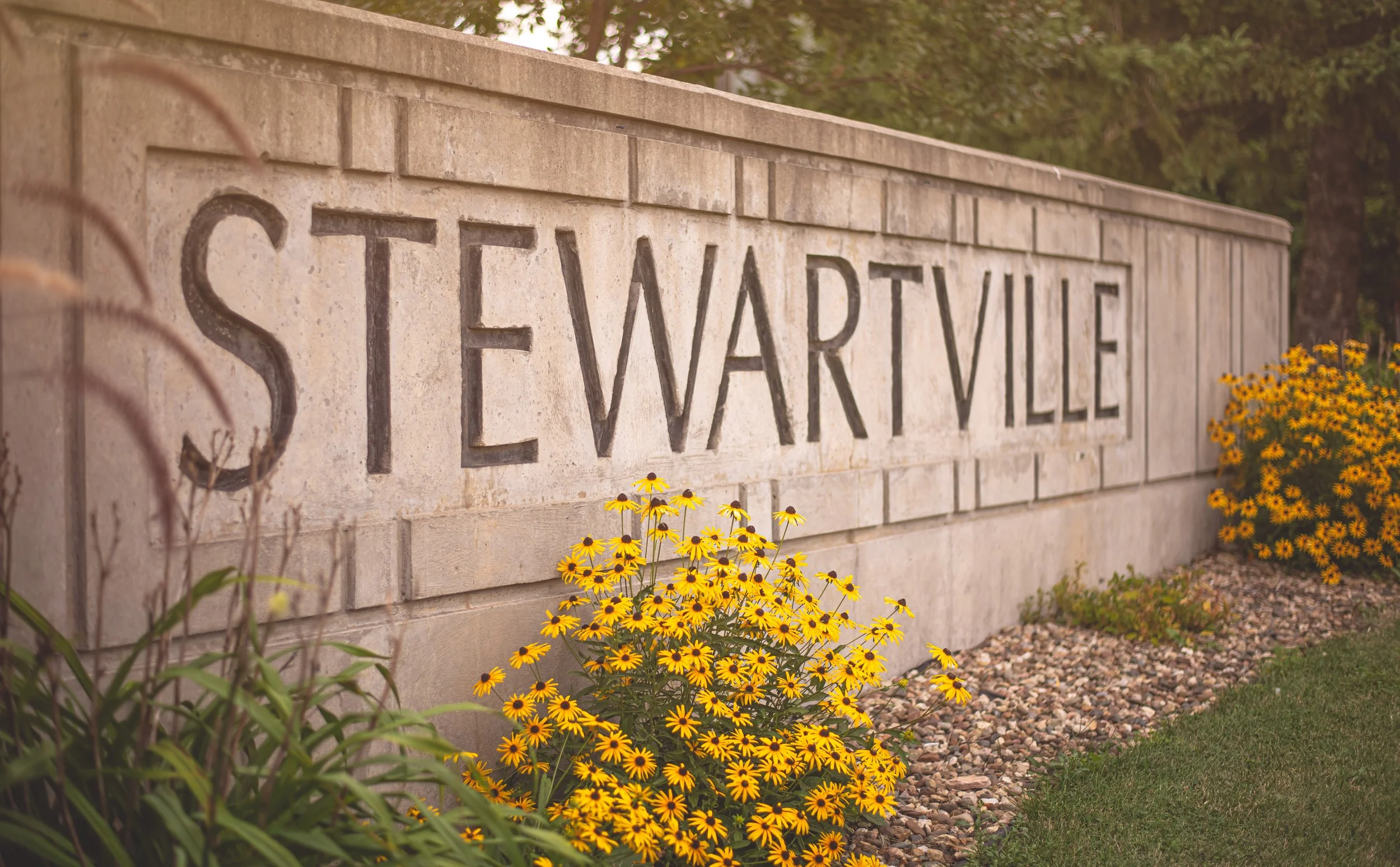 Stone sign with the text 'STEWARTVILLE' engraved on it, surrounded by yellow flowers and greenery.