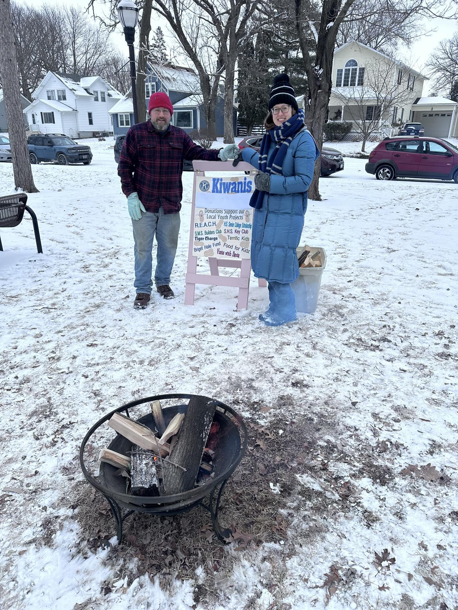 Two people standing outdoors in snowy weather, exchanging a donation box for the Kiwanis club. One person is wearing a red beanie, plaid jacket, and gloves, while the other is wearing a black beanie, blue coat, and glasses. There is a small fire pit 