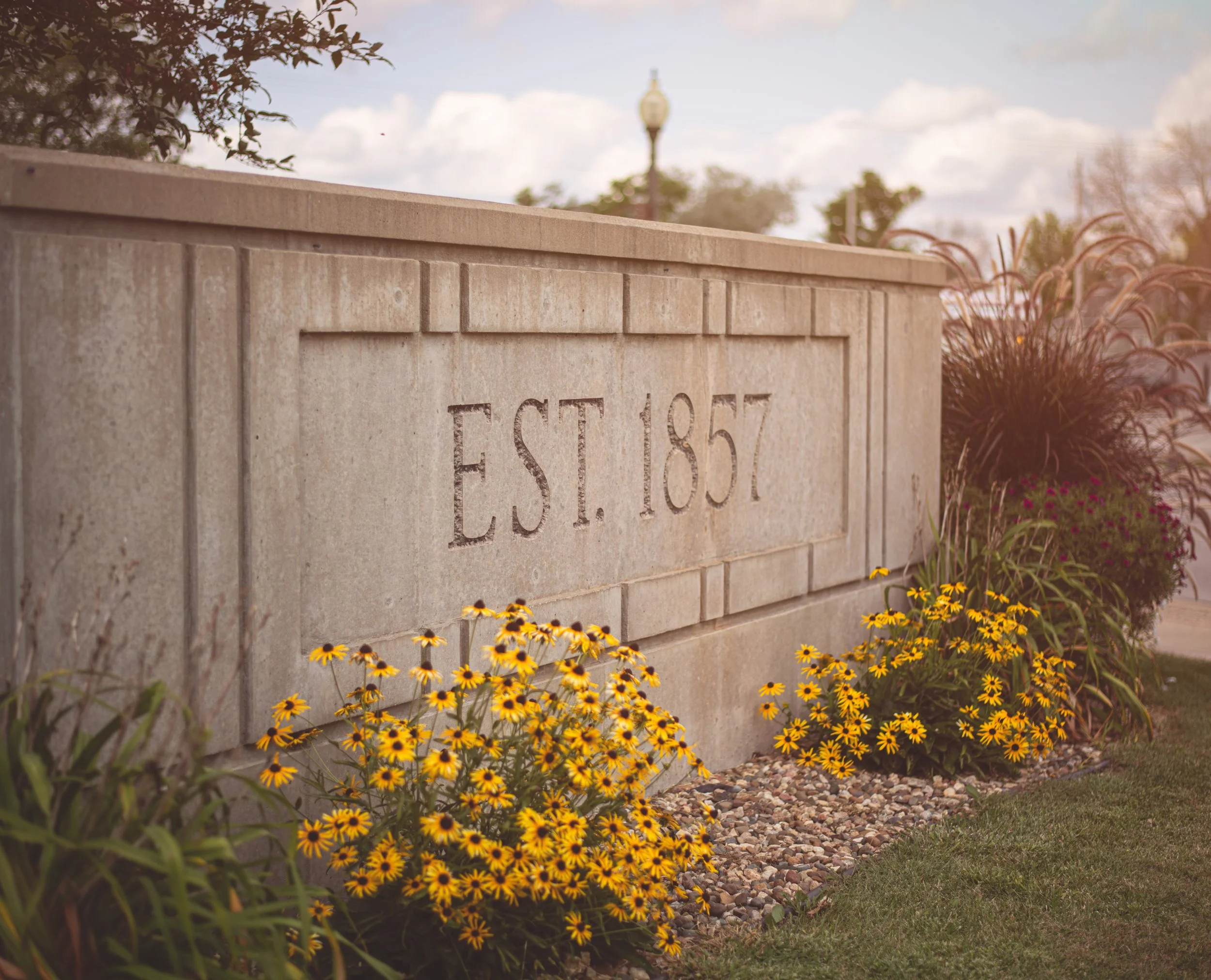 A stone sign with the text 'EST. 1857' engraved on it, surrounded by yellow and purple flowers and greenery, under a partly cloudy sky.