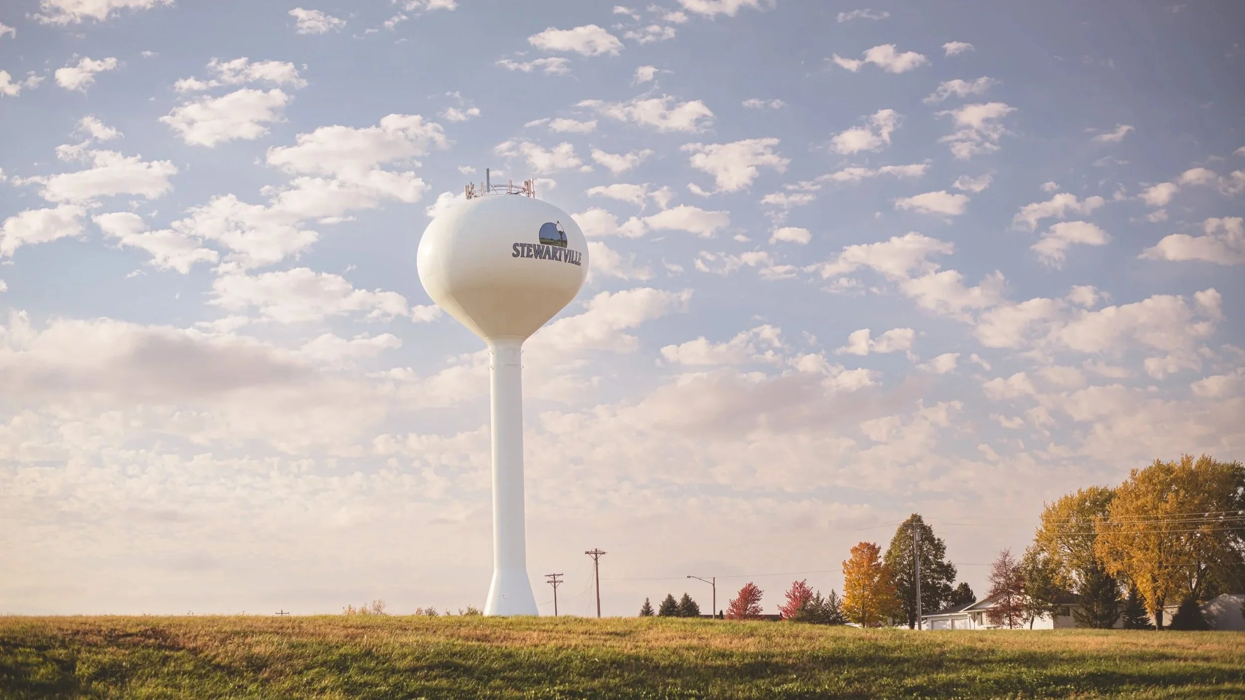 A white water tower with the text 'Stewartville' on it, set against a partly cloudy sky with trees and houses in the background.