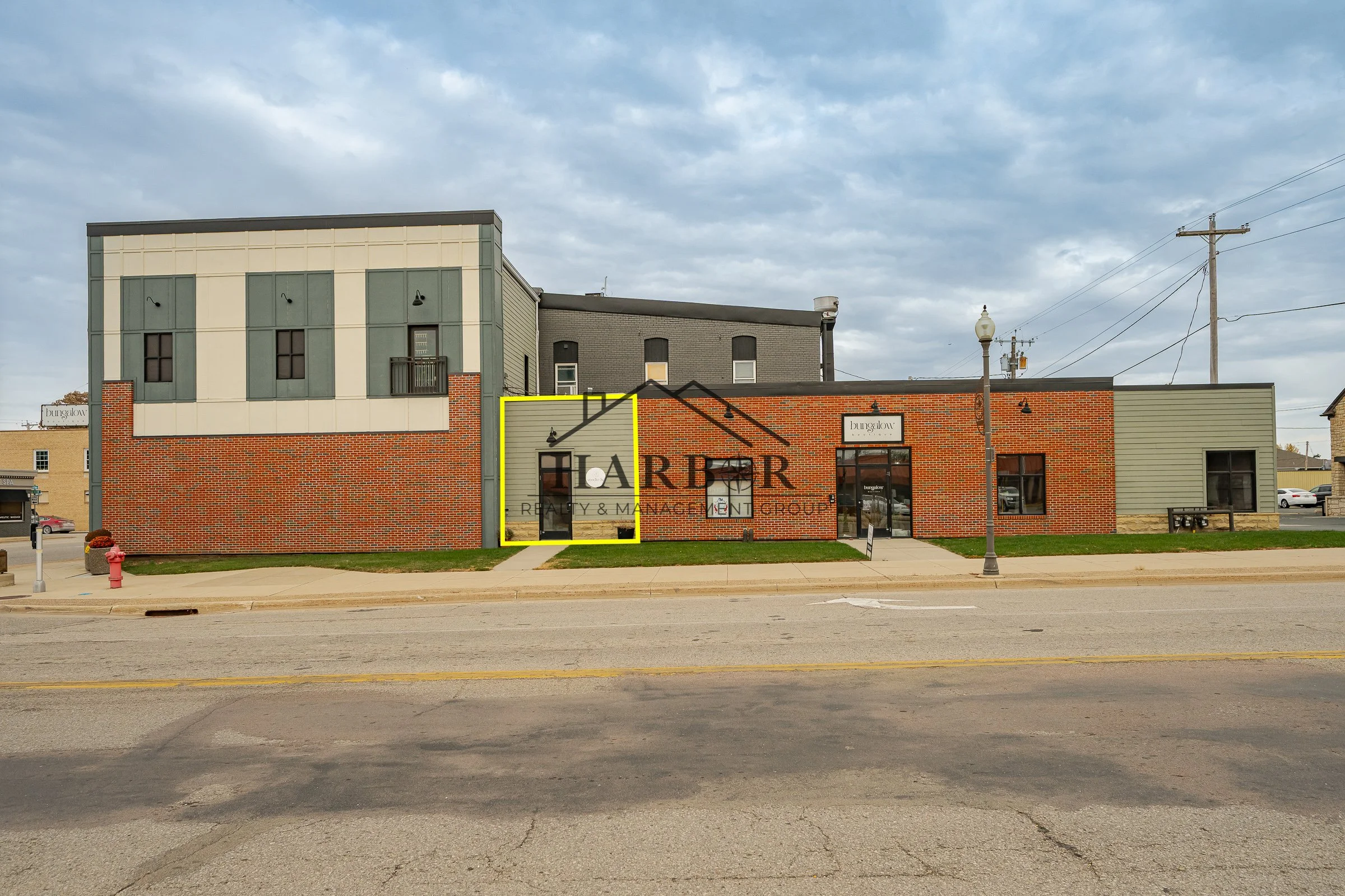 A small commercial building with brick and gray siding, featuring large windows and a black door. A sidewalk runs along the front, with a streetlamp and a 'No Parking' sign nearby. The scene is overcast.