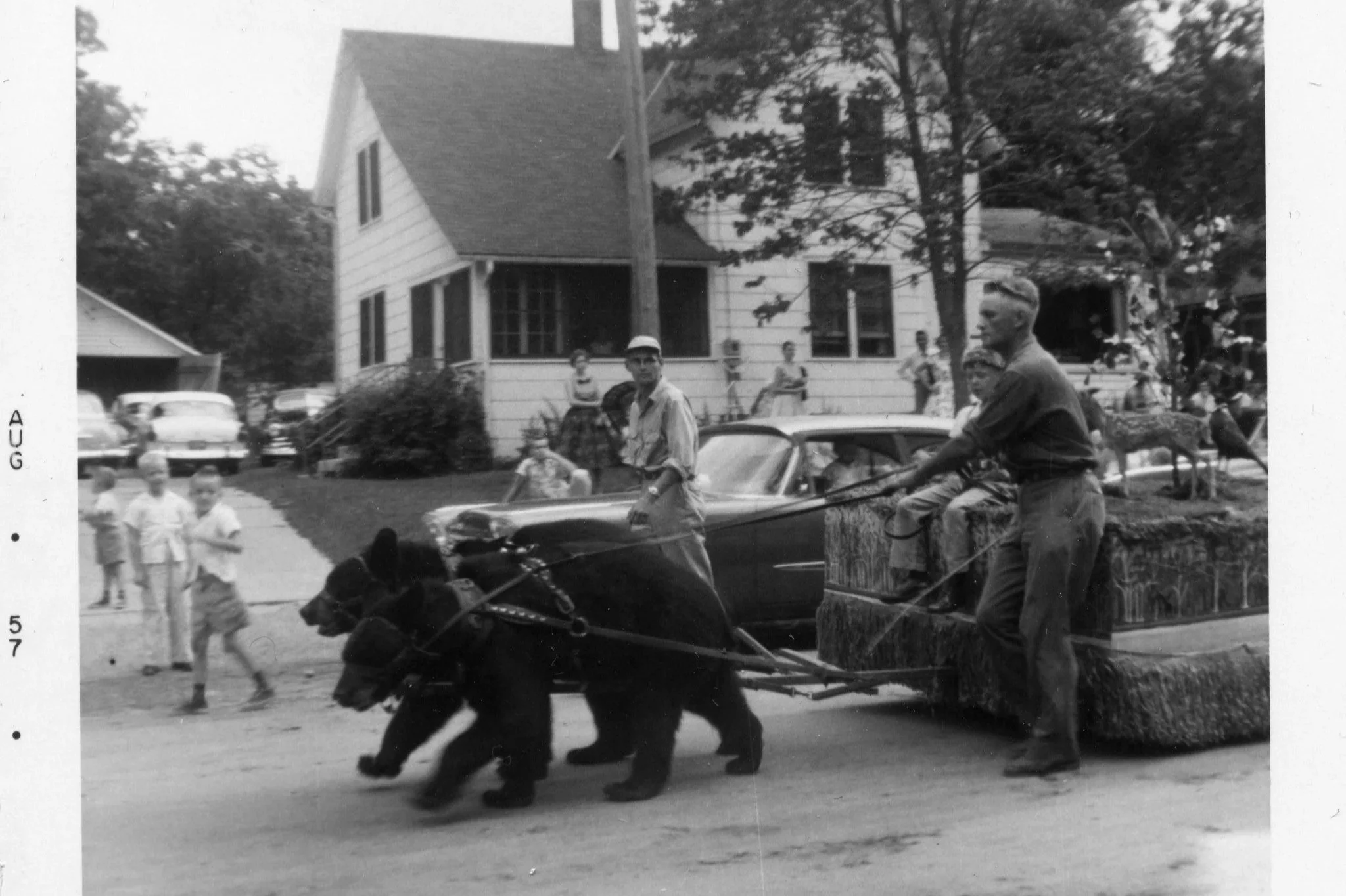 Vintage black and white photograph of children and adults on a street, with a man riding a horse-drawn wagon, and a two-story house in the background.