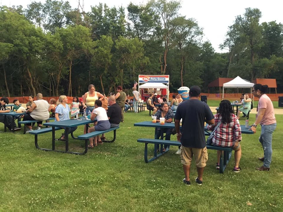 People sitting and standing around blue picnic tables in a grassy park, with trees in the background and food stands in the distance, during an outdoor event.