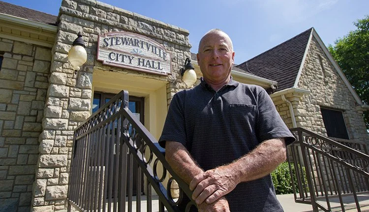 A man standing in front of Stewartville City Hall, a stone building with a sign above the door, decorative exterior lights, and a metal railing on the stairs, under a clear blue sky.