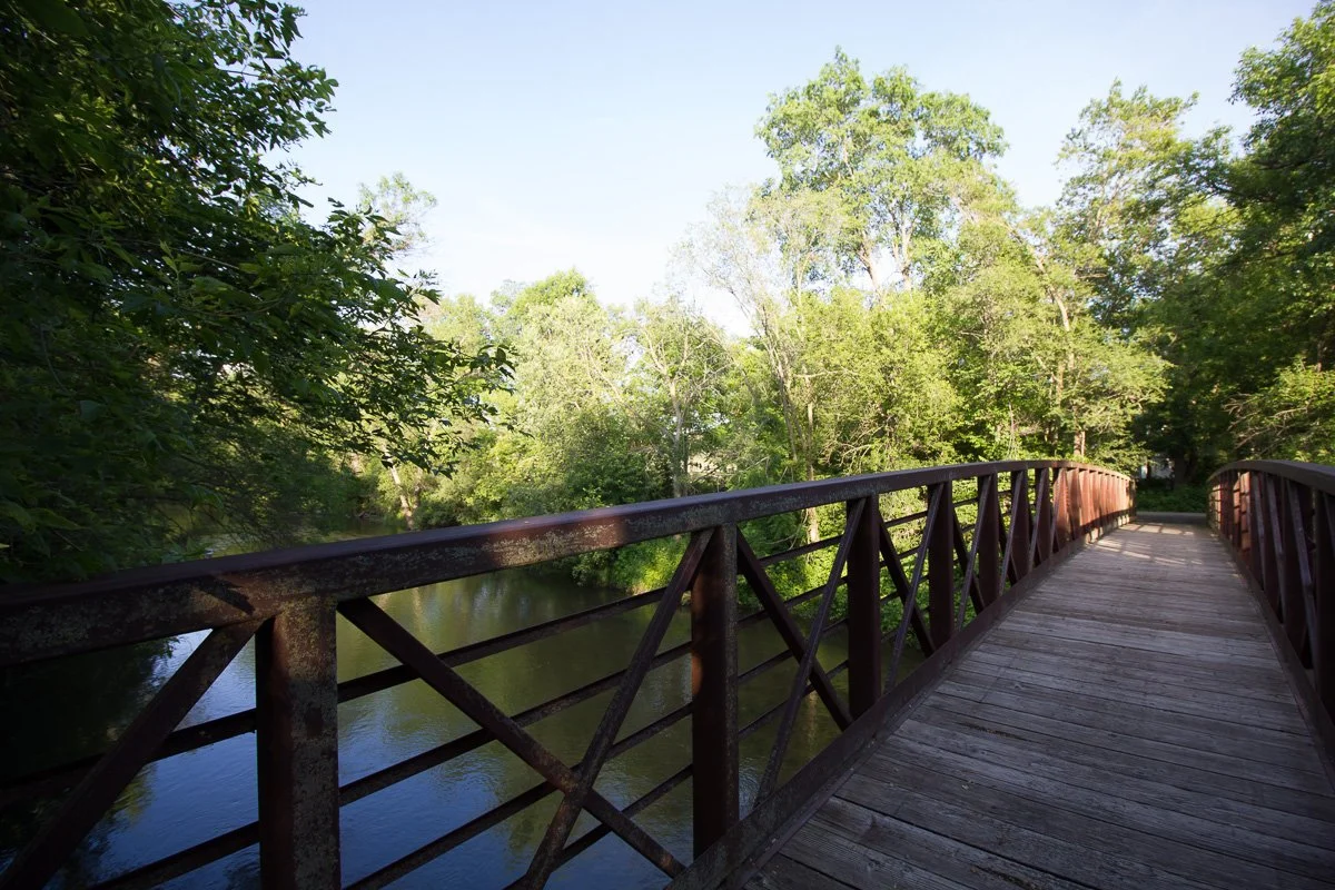 A wooden footbridge with metal railings over a river, surrounded by green trees under a clear sky.