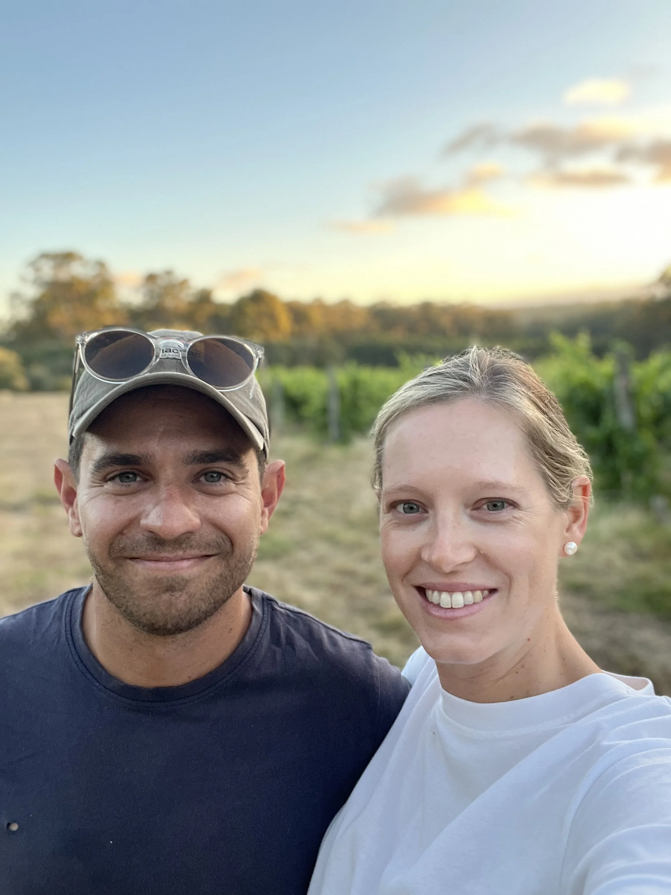A smiling man and woman taking a selfie outdoors during sunset, with a vineyard and trees in the background.