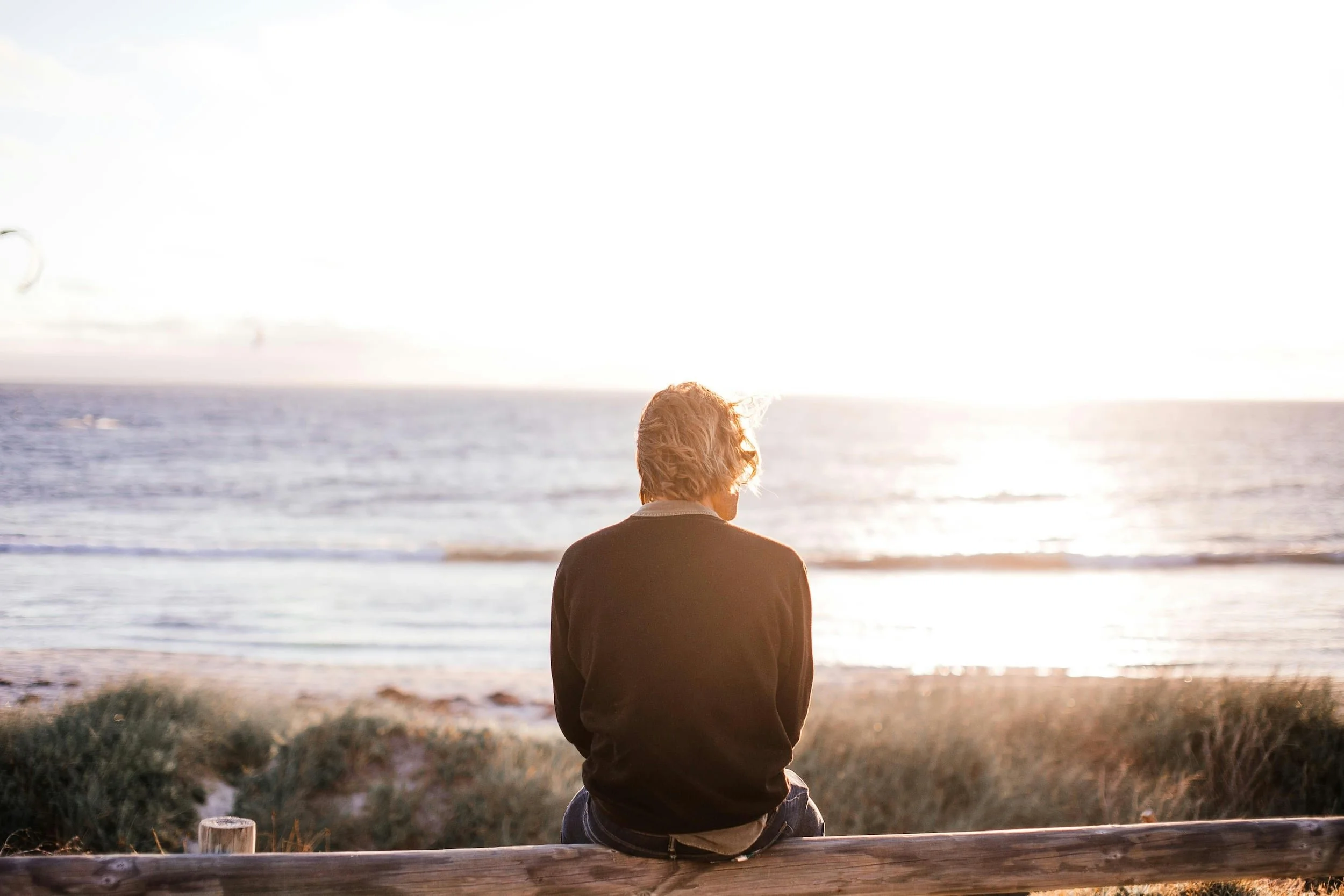 Person sitting alone on the beach, gazing toward the horizon under soft light, creating a quiet and reflective mood.