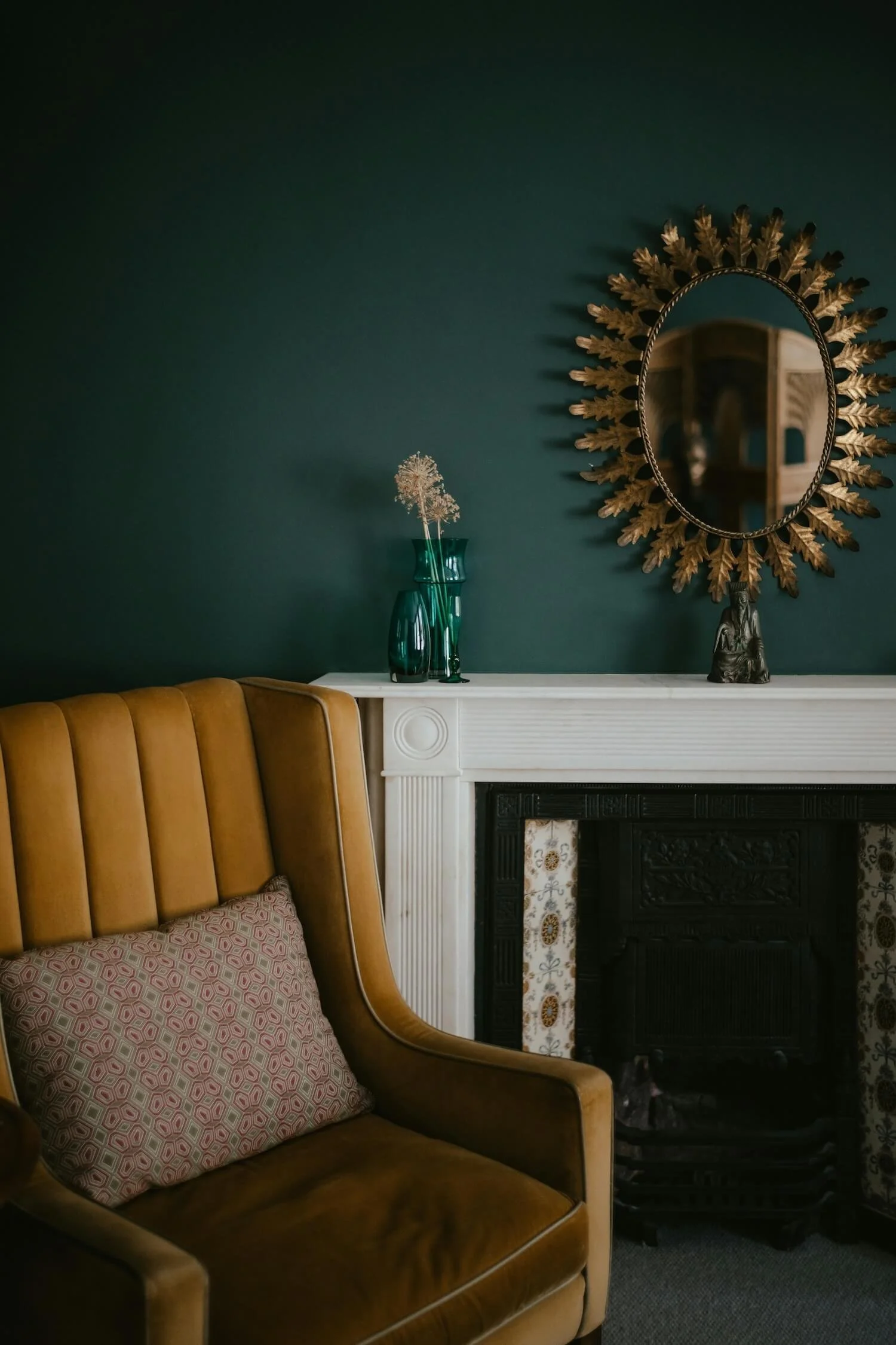 Cosy room with a mustard armchair beside a white fireplace and round mirror on a dark green wall.