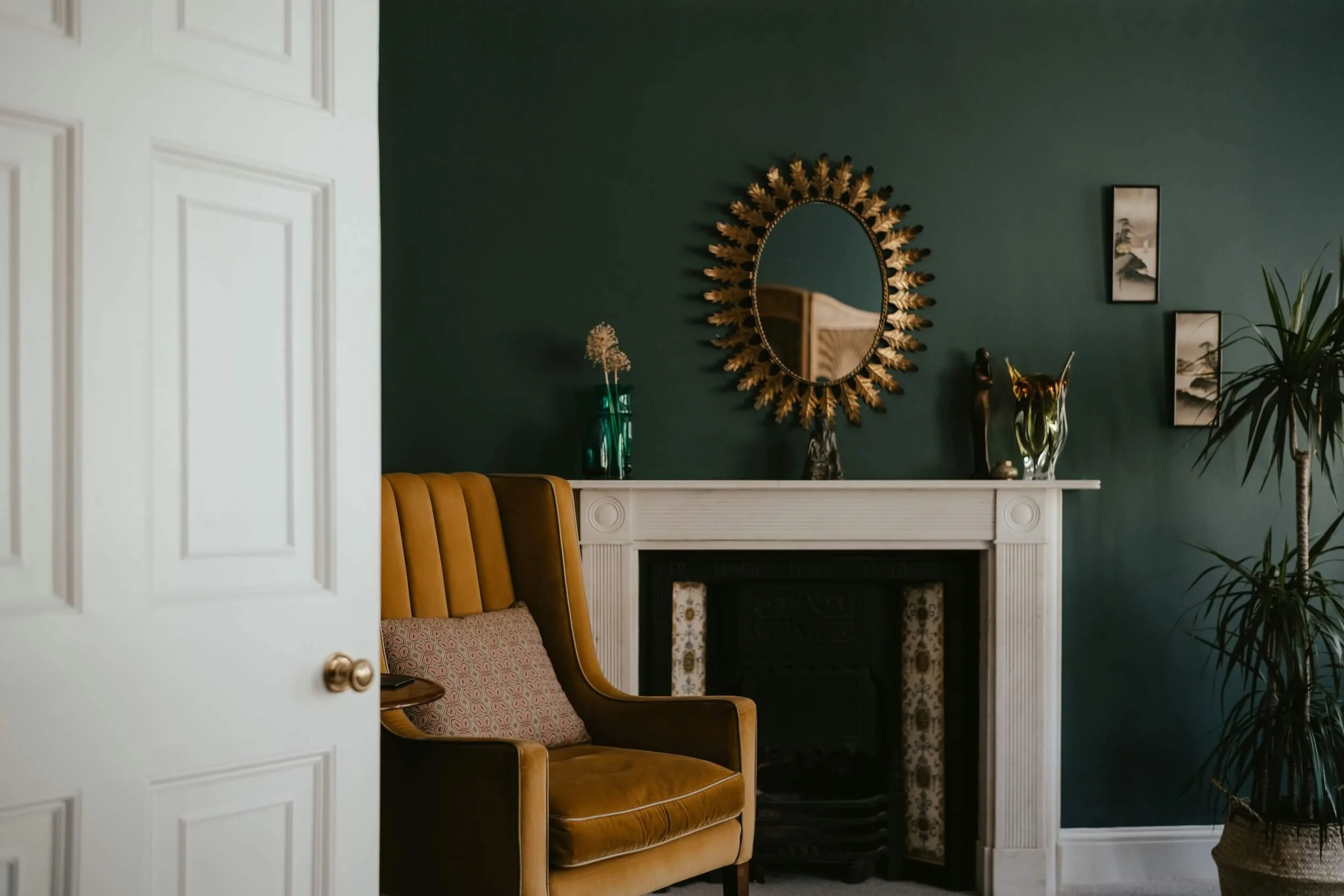 Interior of a calm, stylish therapy room featuring a mustard armchair beside a white fireplace, with a round mirror and plant on a dark green wall.