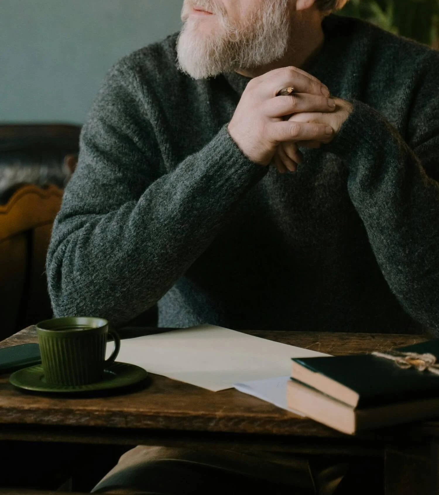 Man in a dark sweater sitting at a wooden desk with a cup of tea and open notebook, reflecting quietly.