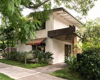 A two-story white house with a dark awning over the front entrance, surrounded by green trees and plants, with a sidewalk running in front.