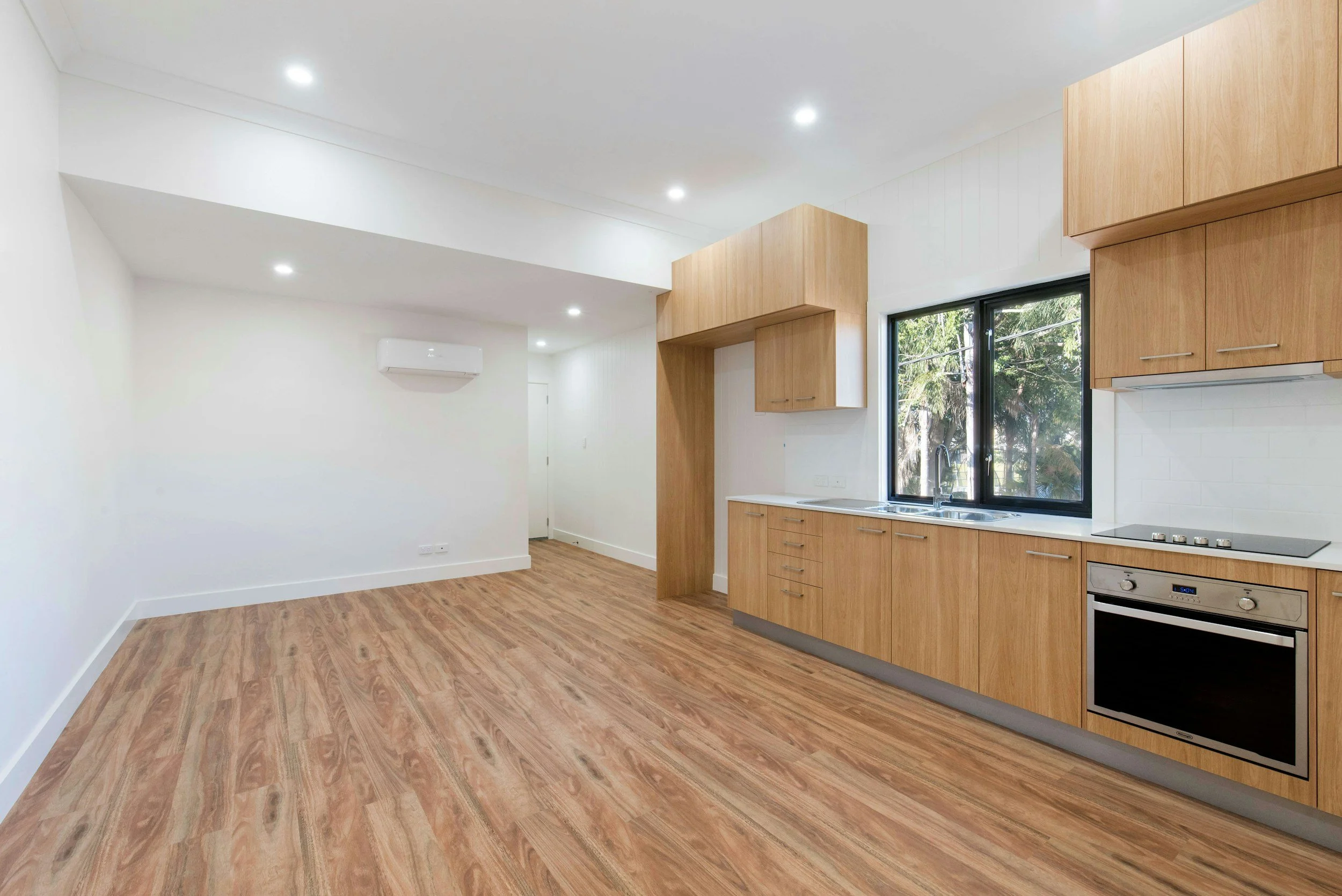 Empty kitchen with wooden cabinets, a window showing trees outside, and built-in appliances including an oven and an electric cooktop, with white walls and wood flooring.